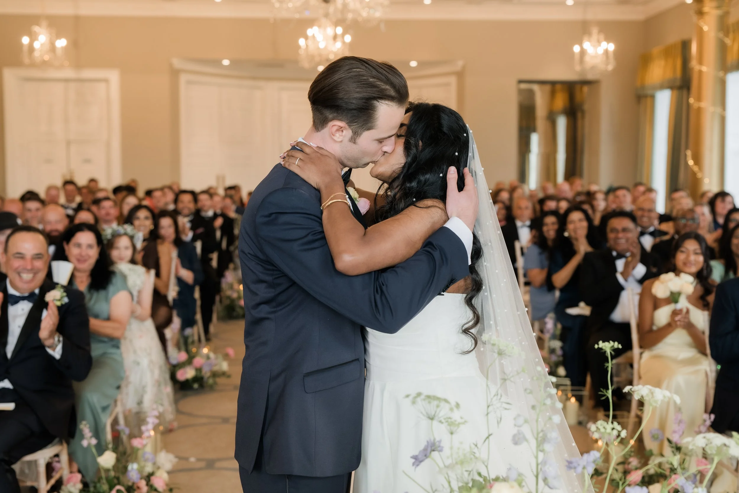 A couple shares a kiss during their wedding ceremony in a decorated indoor venue filled with seated guests.