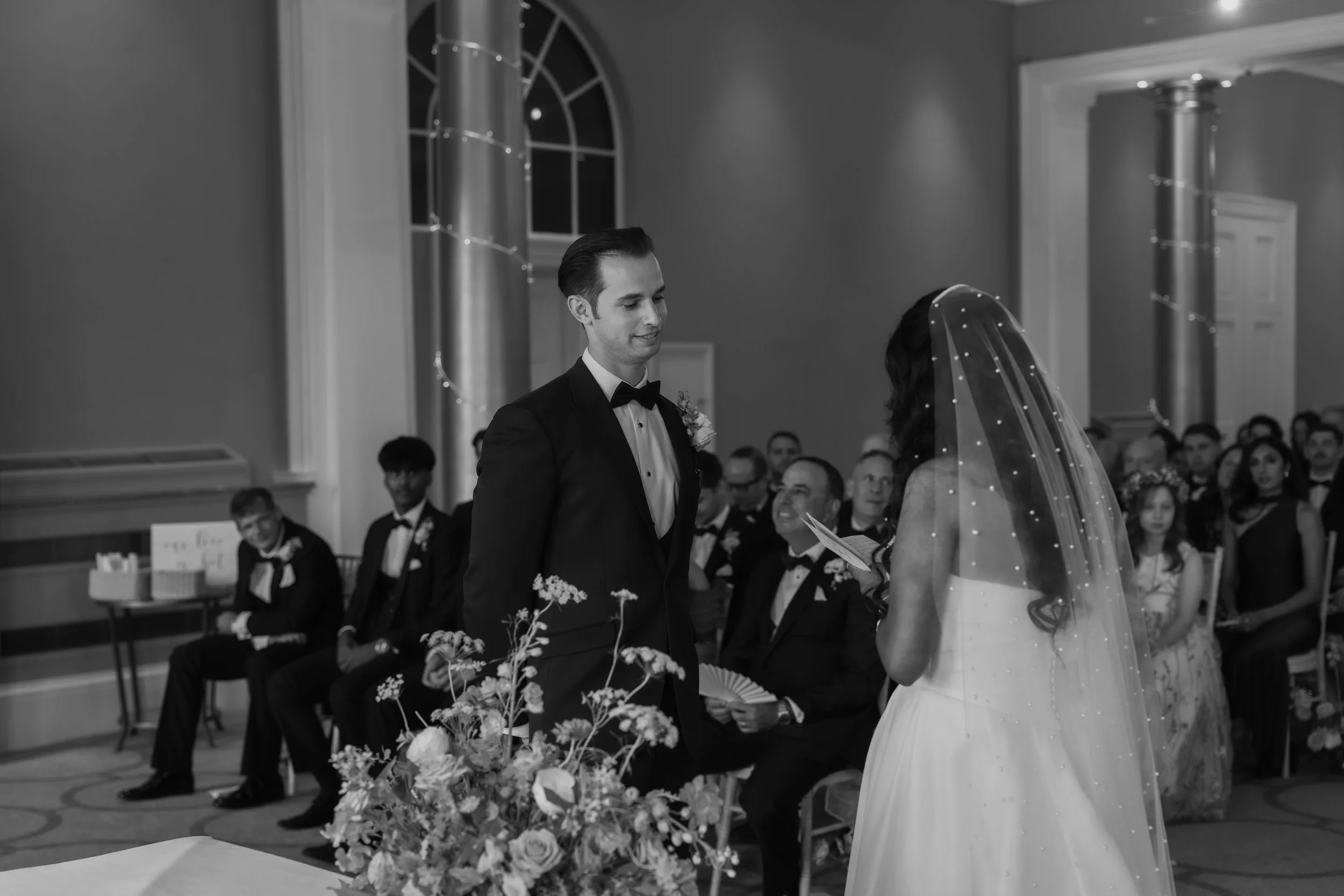 A black-and-white photo of a wedding ceremony, with a groom and bride standing in front of each other, focusing on each other's faces. The groom wears a tuxedo and the bride wears a wedding dress with a veil. Guests are seated in the background, smil