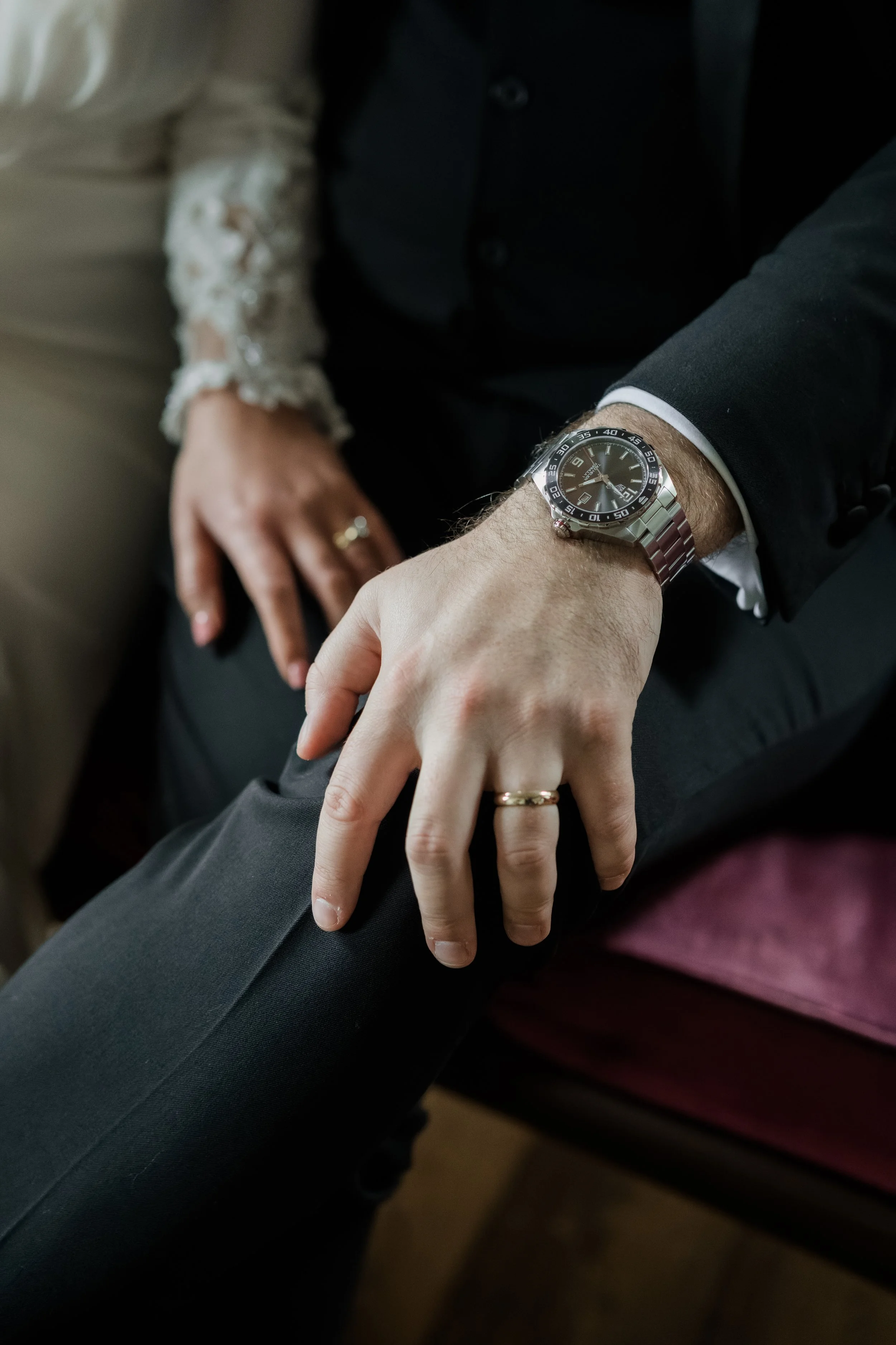 Close-up of a man and woman holding hands, the man wearing a watch and wedding band, sitting on a sofa.