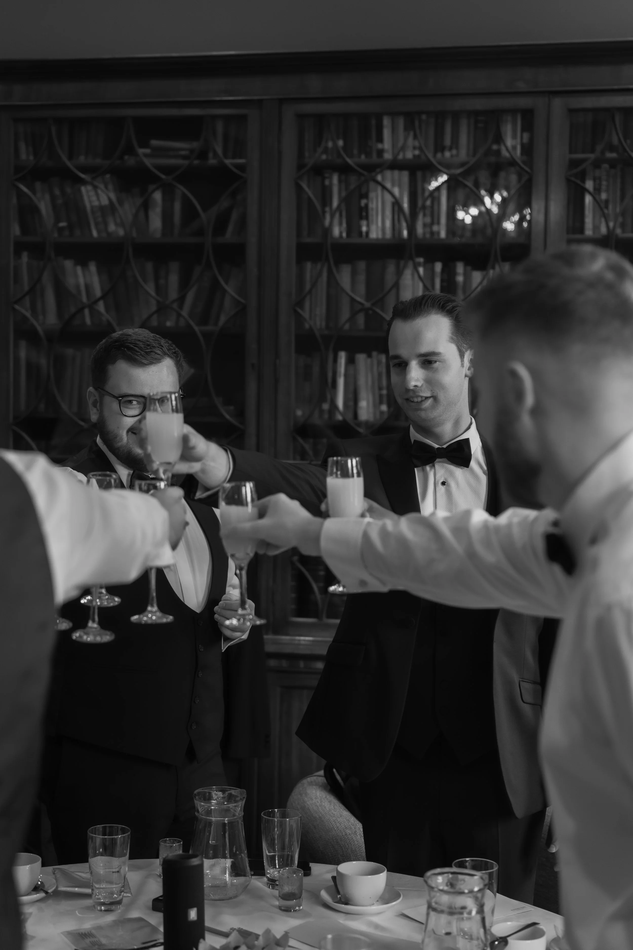 Four men, dressed in tuxedos, raise glasses in a toast at a formal gathering in a room with a wooden bookcase filled with books in the background.