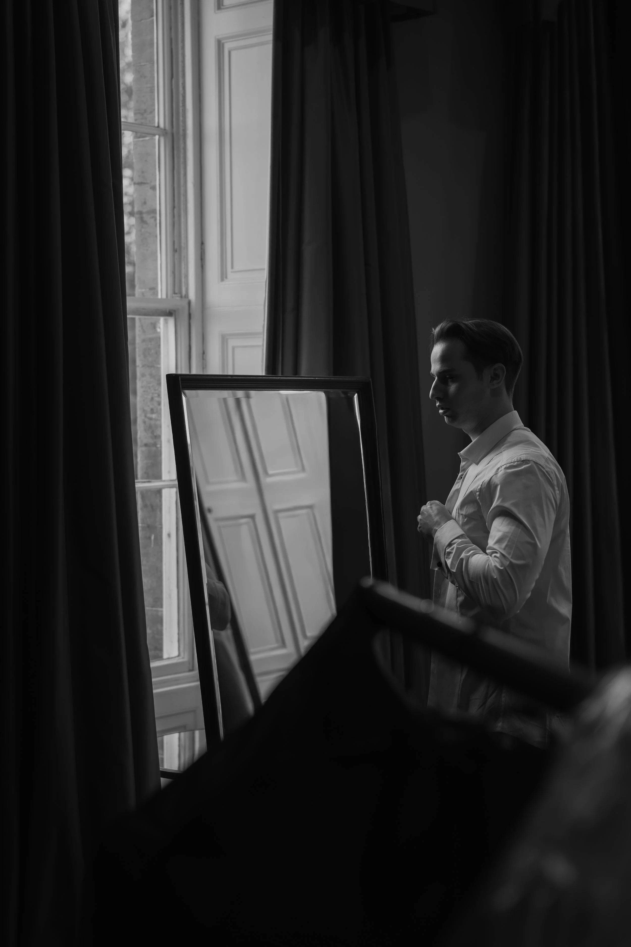 A man in a white shirt looking at himself in a mirror, with a large window and curtains in the background, in a black and white photograph.