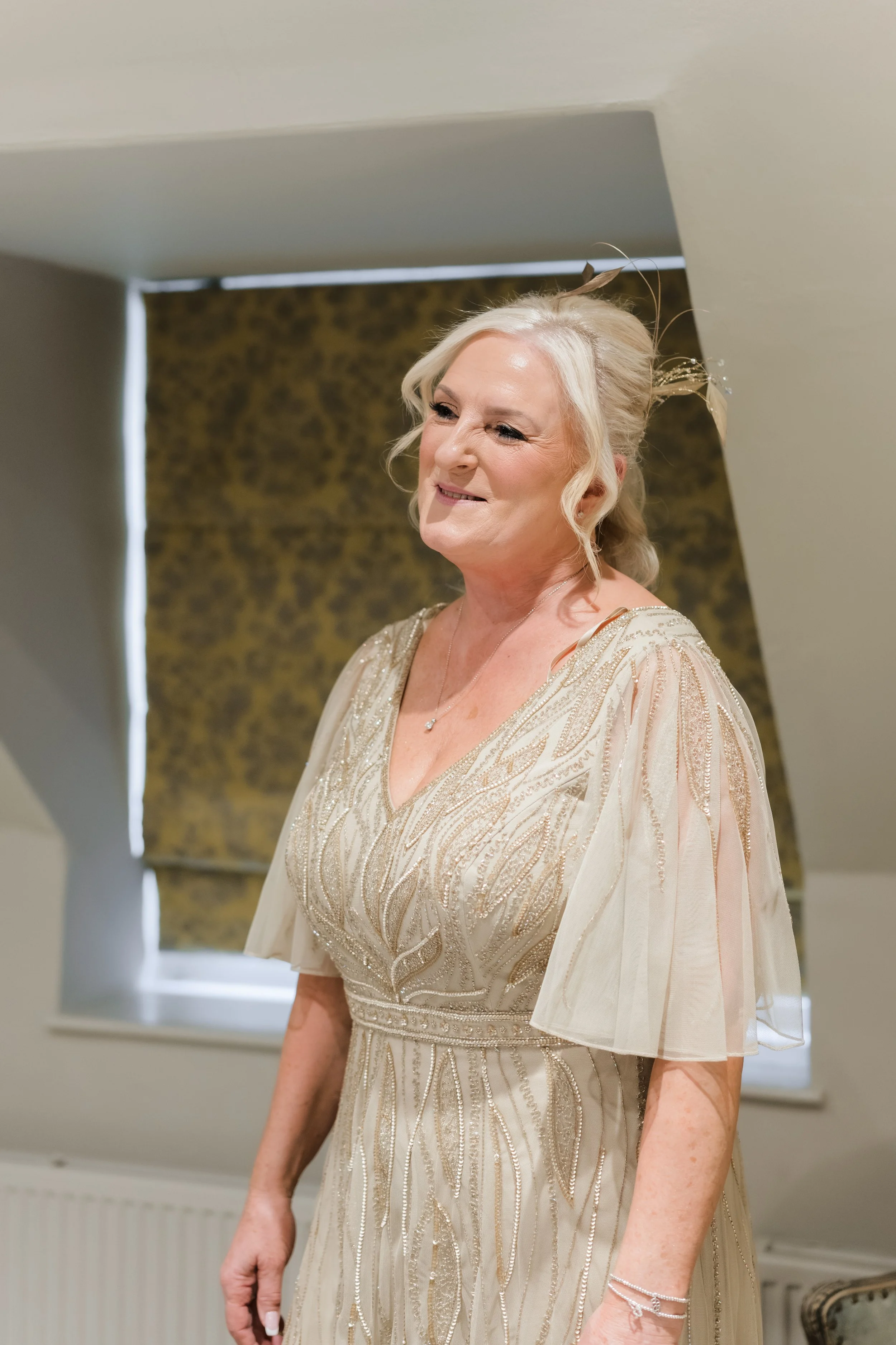 A smiling older woman in a beige, embroidered dress, standing indoors near a window with a closed, patterned blind.