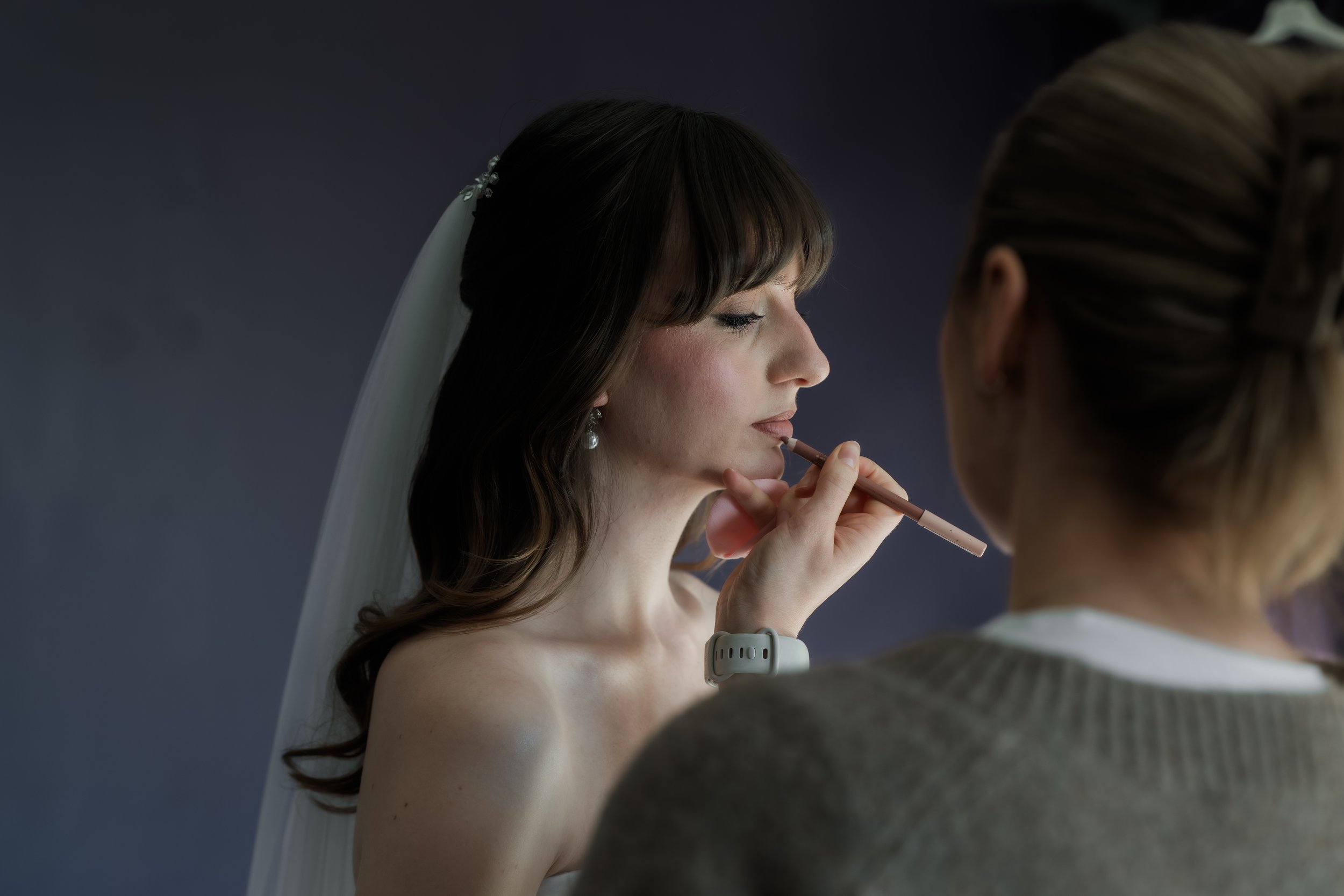 A bride in a wedding dress has makeup applied by a makeup artist.