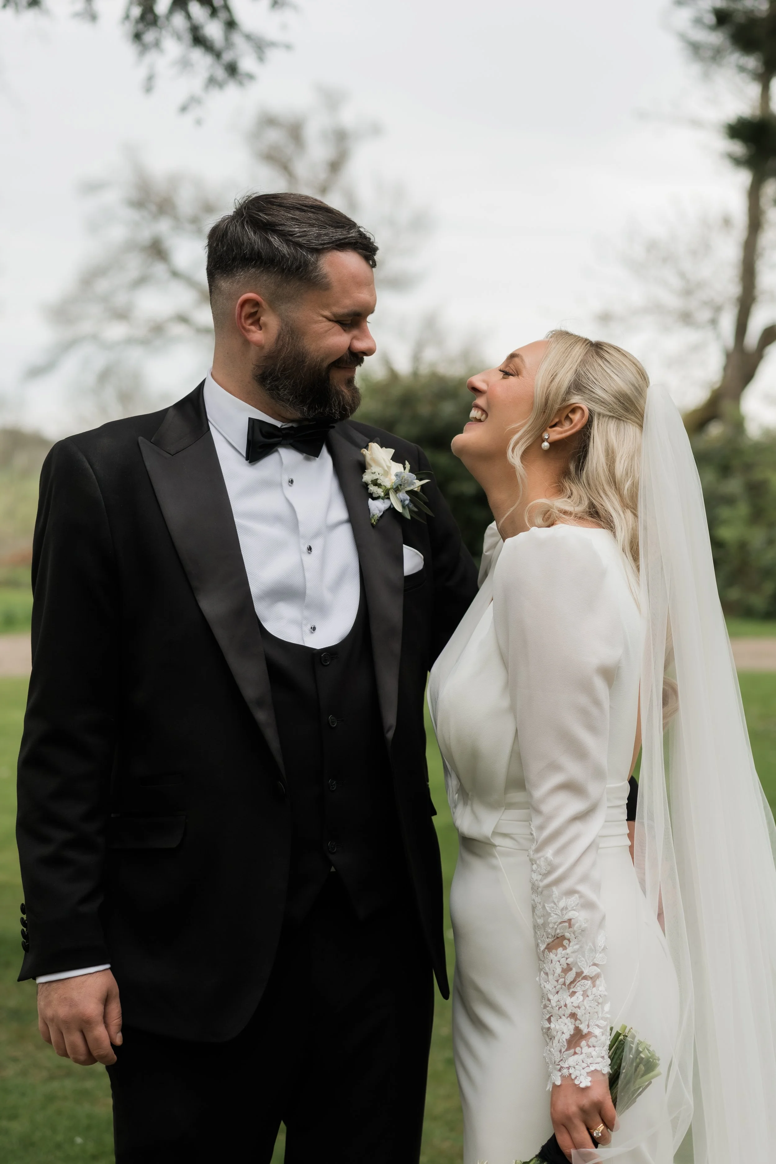 A bride and groom smiling and looking at each other outdoors on their wedding day. The groom is wearing a black tuxedo with a bow tie, and the bride is in a white wedding dress with lace details, holding a small bouquet, with a veil and pearl earring