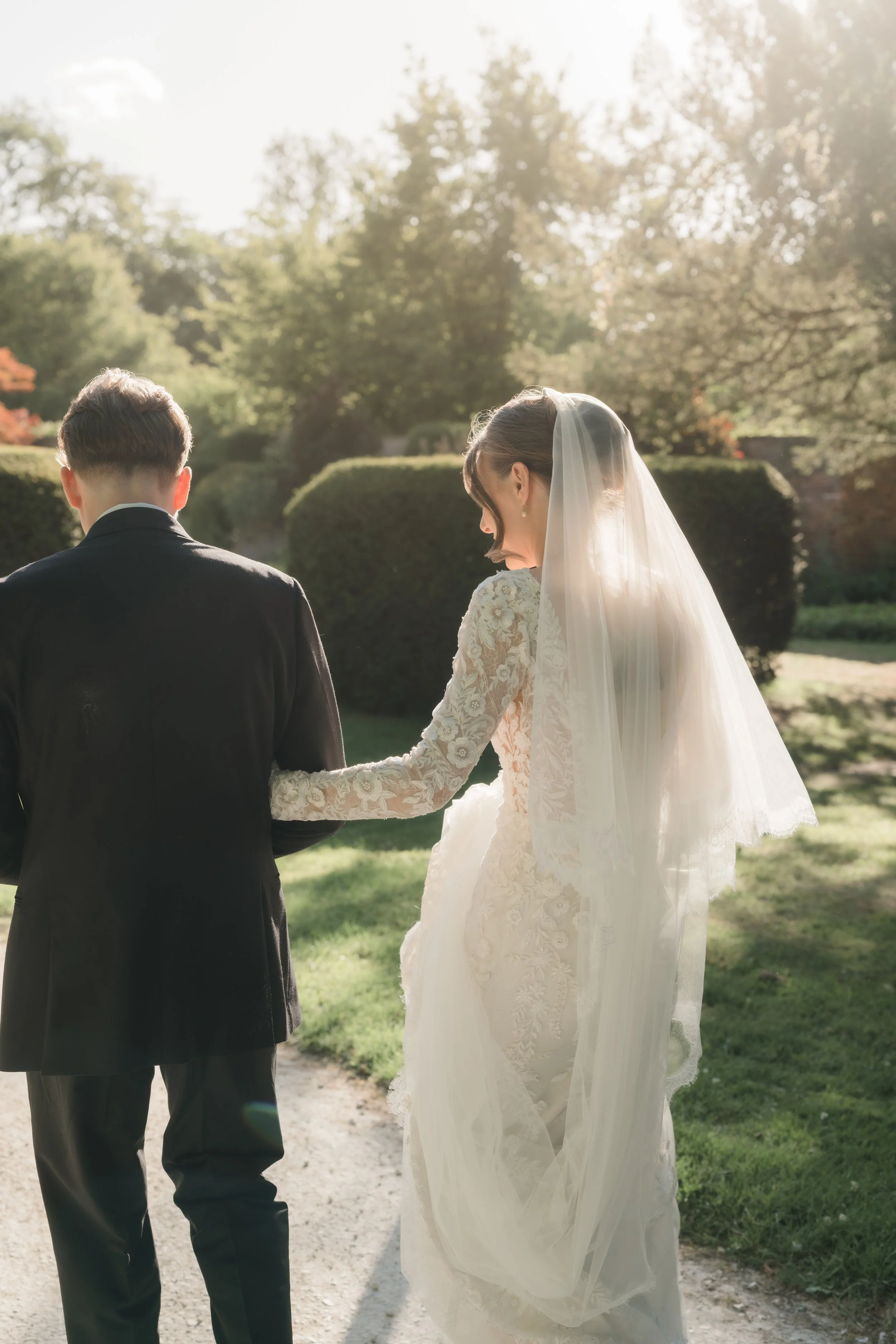 A bride and groom holding hands outdoors during a sunny wedding ceremony. The bride wears a lace wedding dress with a train and a veil, while the groom in a black suit. The background features trees and bushes.