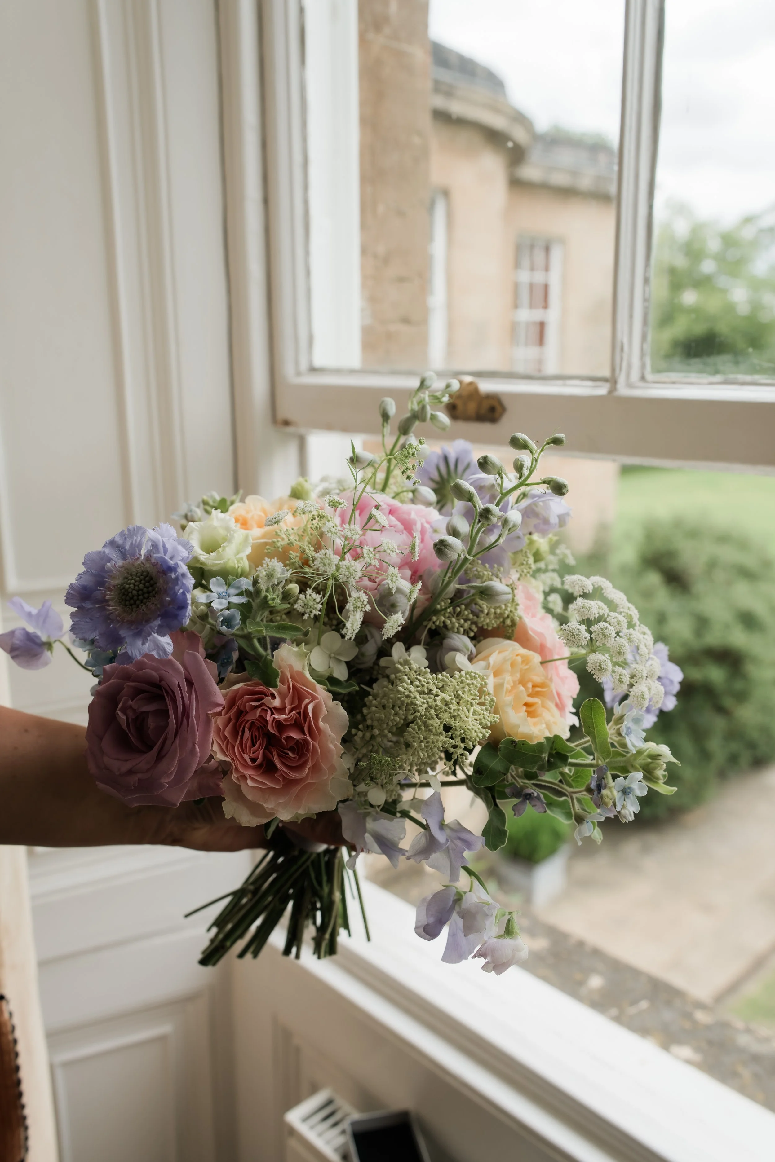 A person holds a colorful bouquet of flowers near a window with a view of the outdoor building and greenery.
