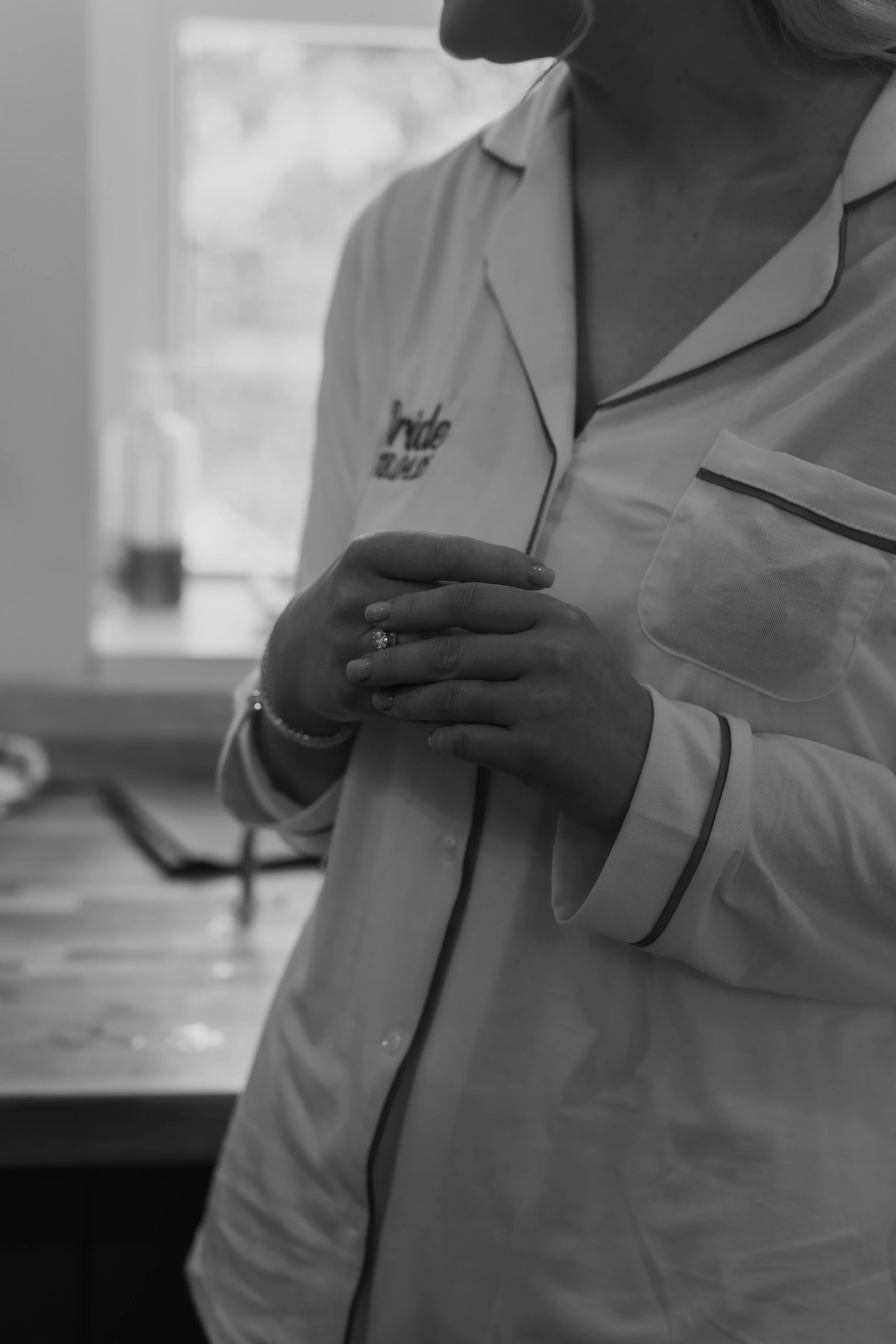 A woman in pajamas, wearing bracelets and a ring, standing indoors with hands clasped, near a window.