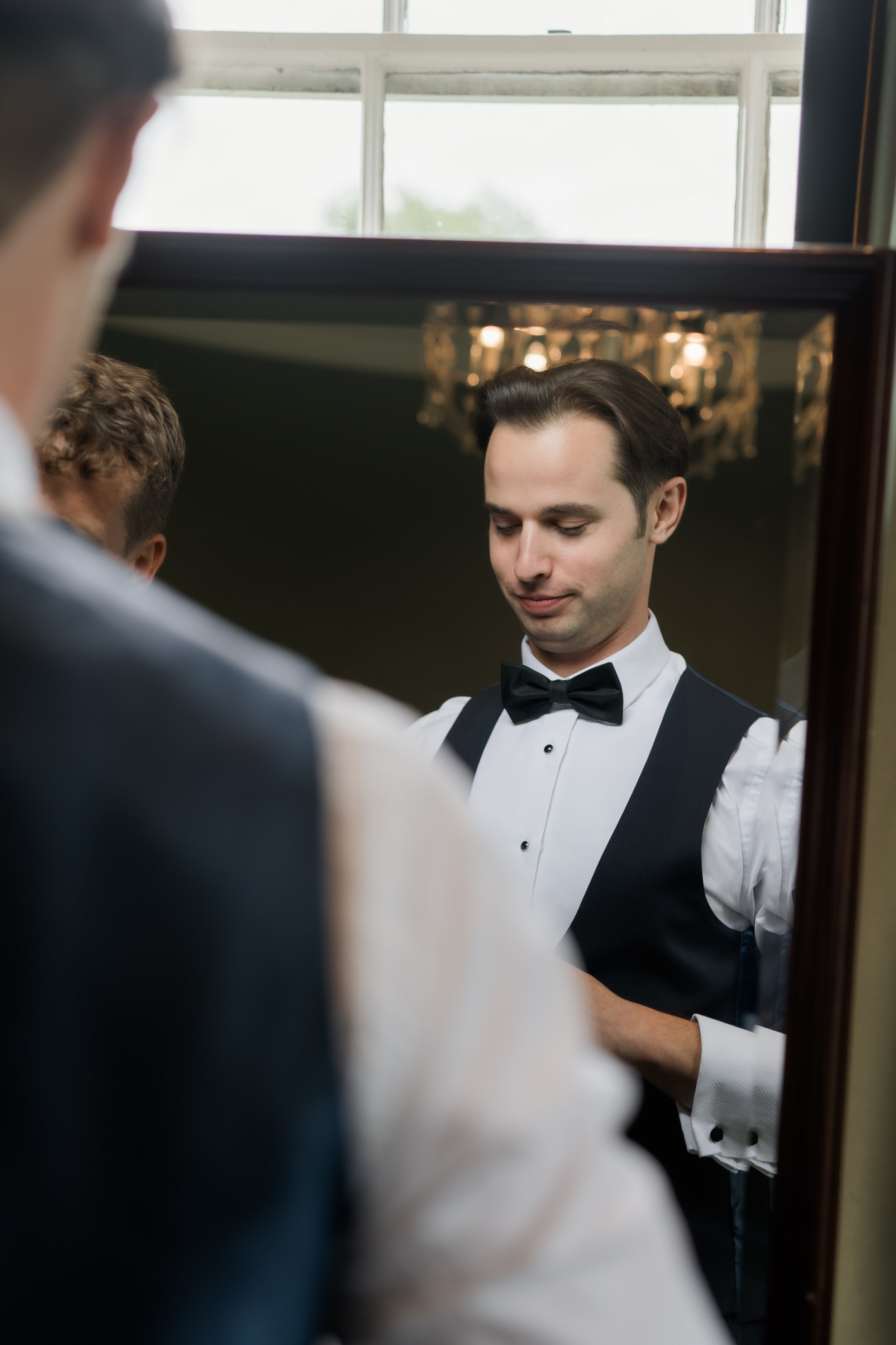 A groom in a tuxedo looking at himself in a mirror, presumably before a wedding.