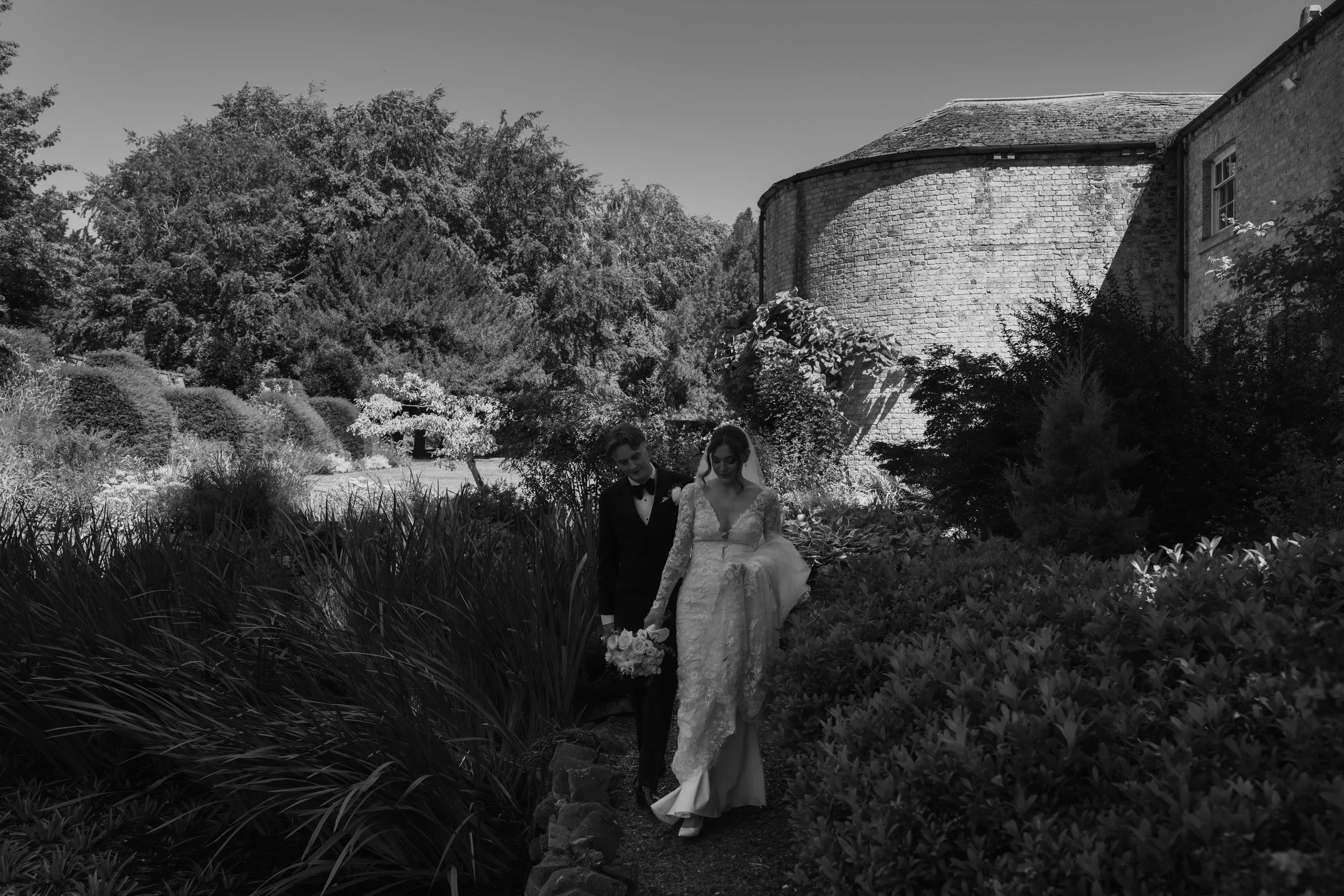 Black and white groom walks behind bride as they walk around Saltmarshe Hall pond 