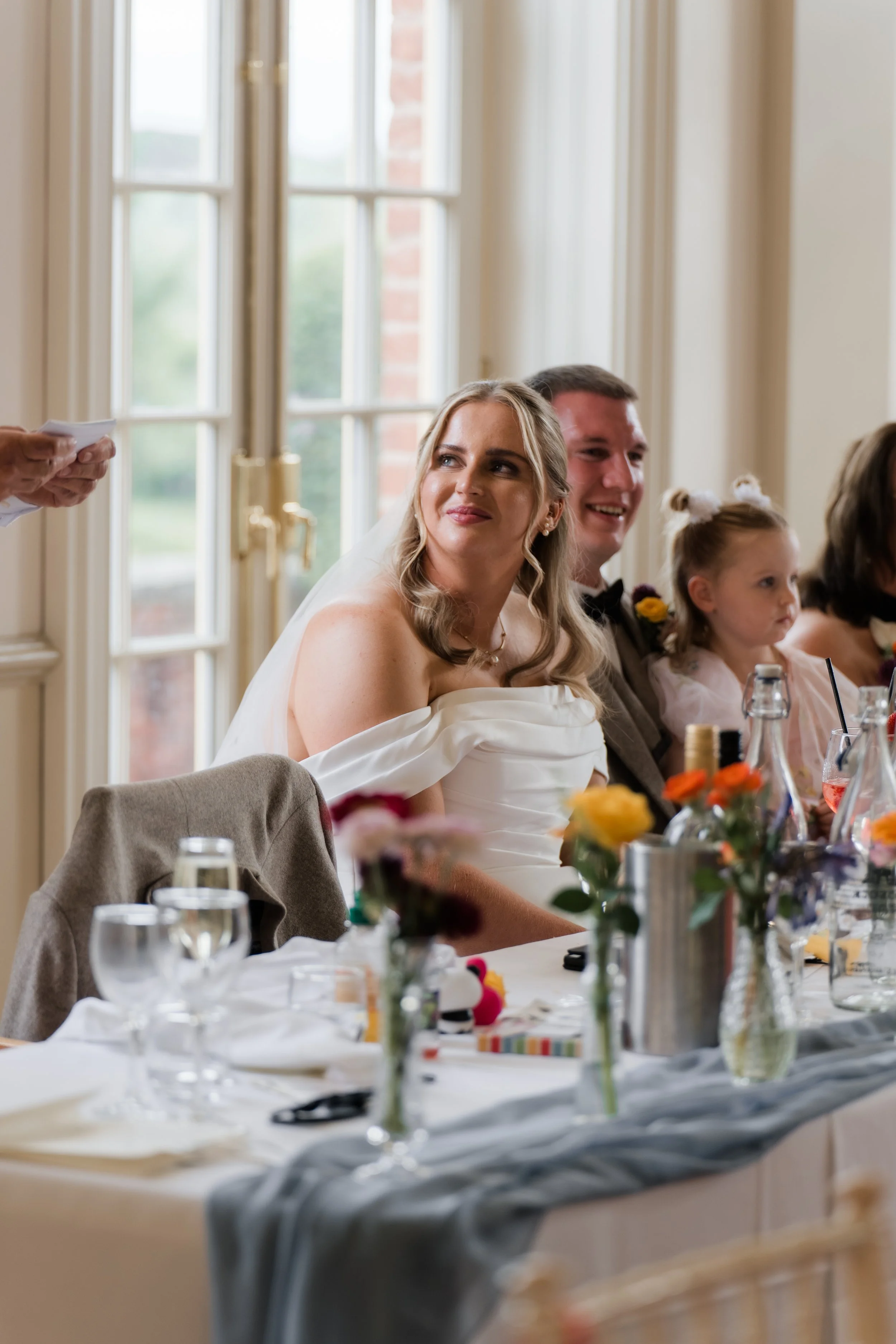 Bride and groom sitting at a wedding reception table, smiling, with floral centerpieces and glassware.