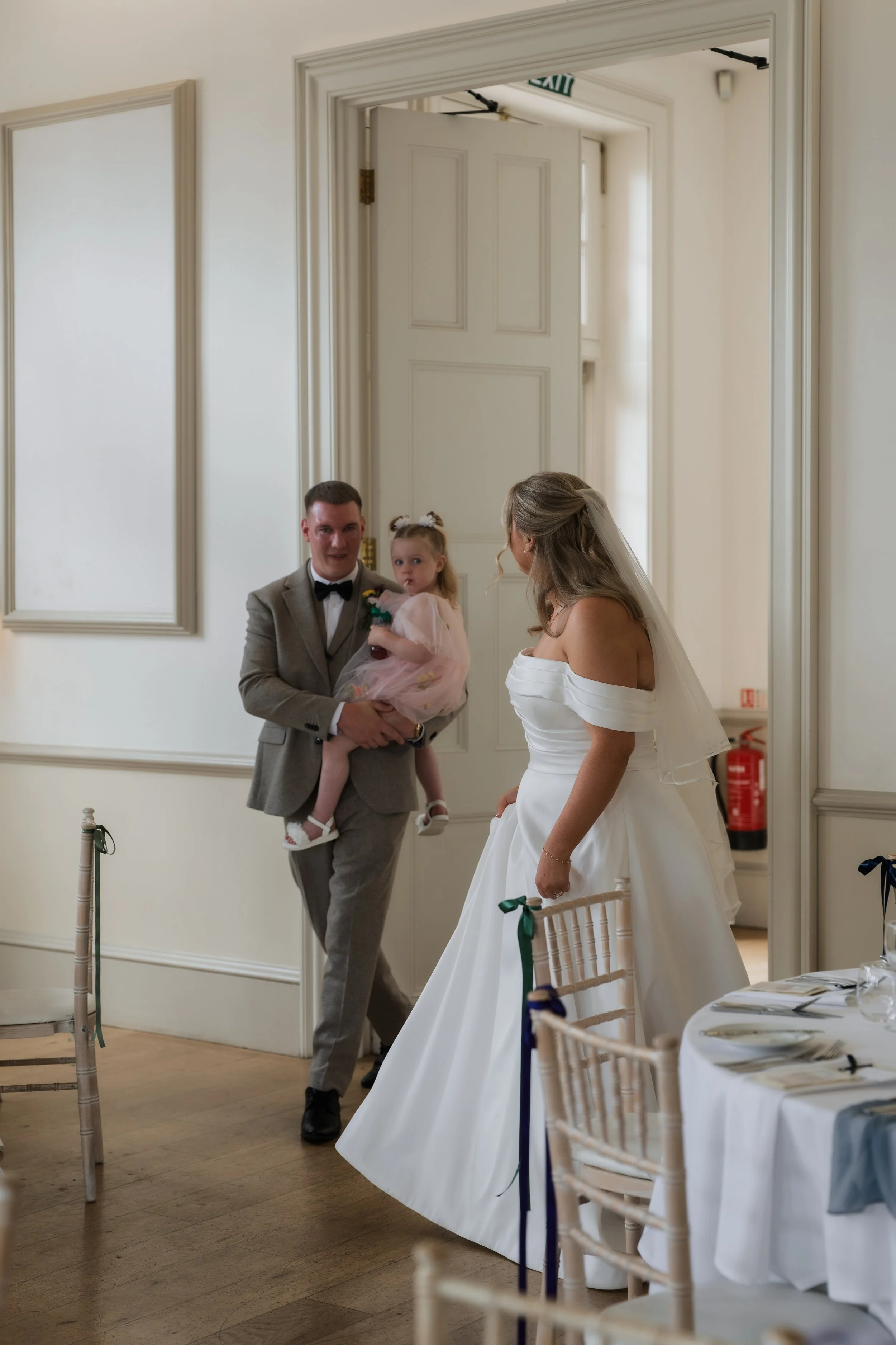 A bride in a white wedding dress interacts with a man in a suit and a young girl in a pink dress in a decorated event room.