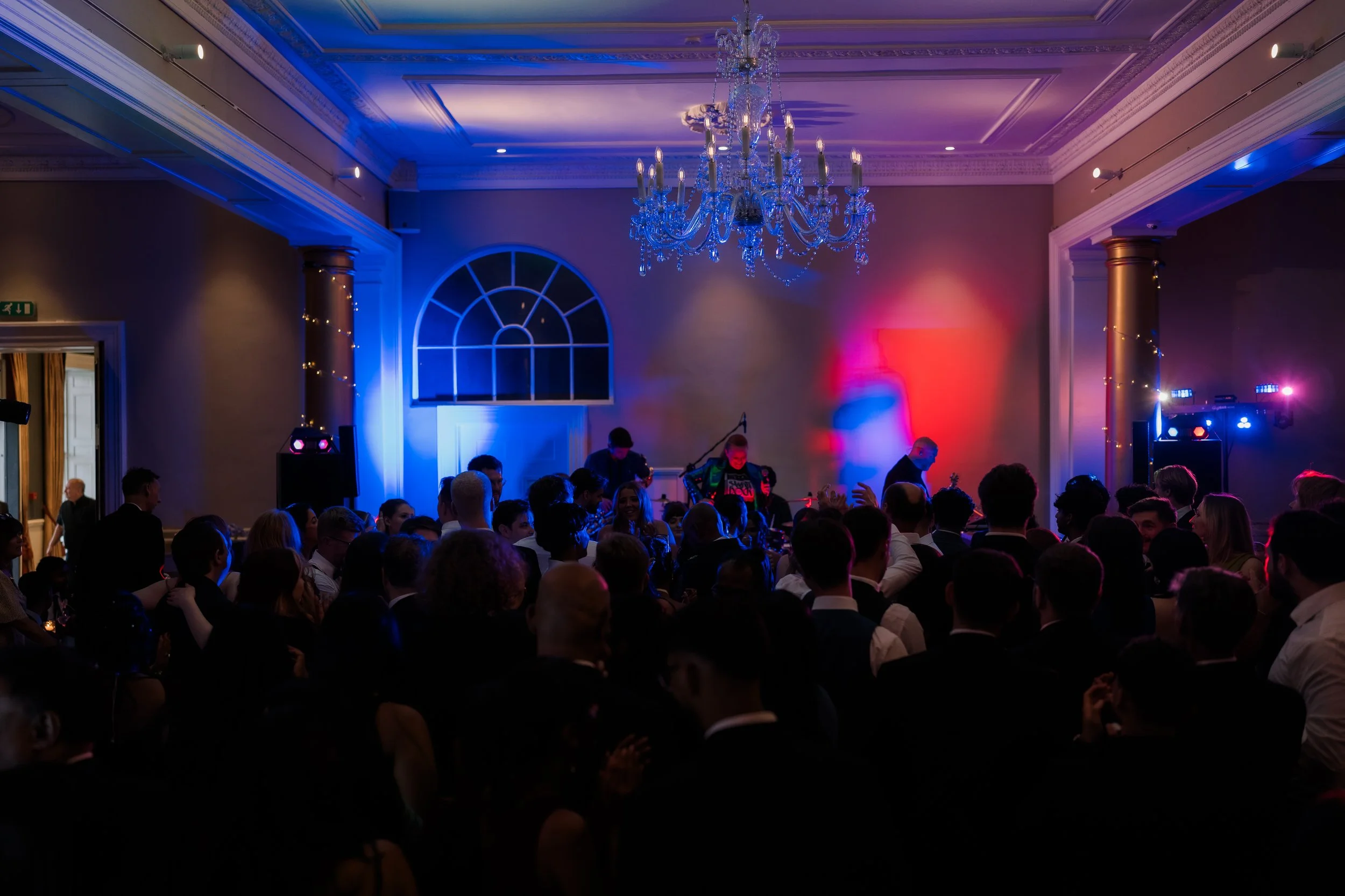 Crowd gathered at a formal event with a live band performing on stage in a large decorated ballroom, illuminated with colorful lights, a chandelier hangs from the ceiling, and people are dressed in formal attire.
