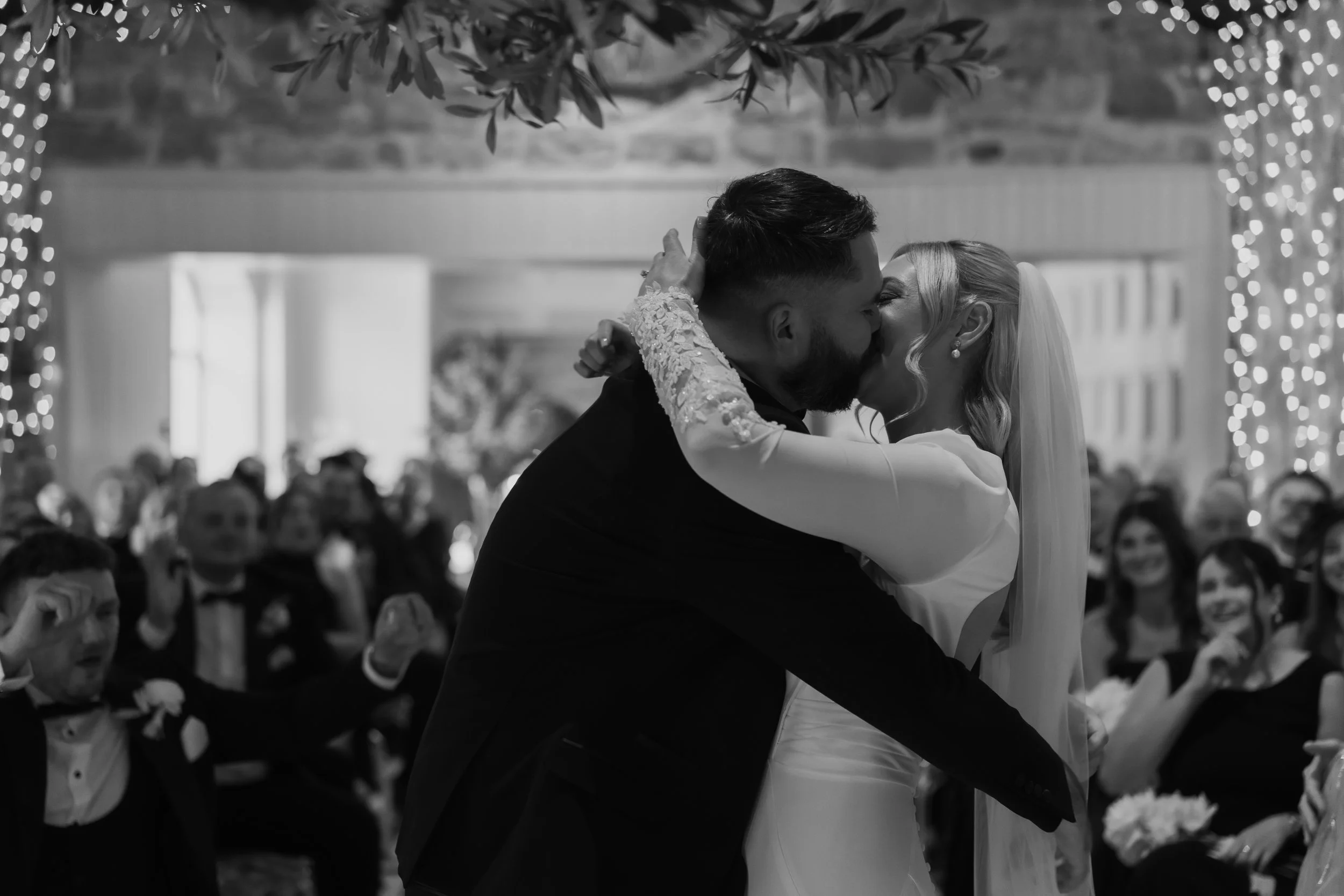 Black and white photograph of a bride and groom sharing a kiss during their wedding ceremony, surrounded by seated guests in an indoor venue.