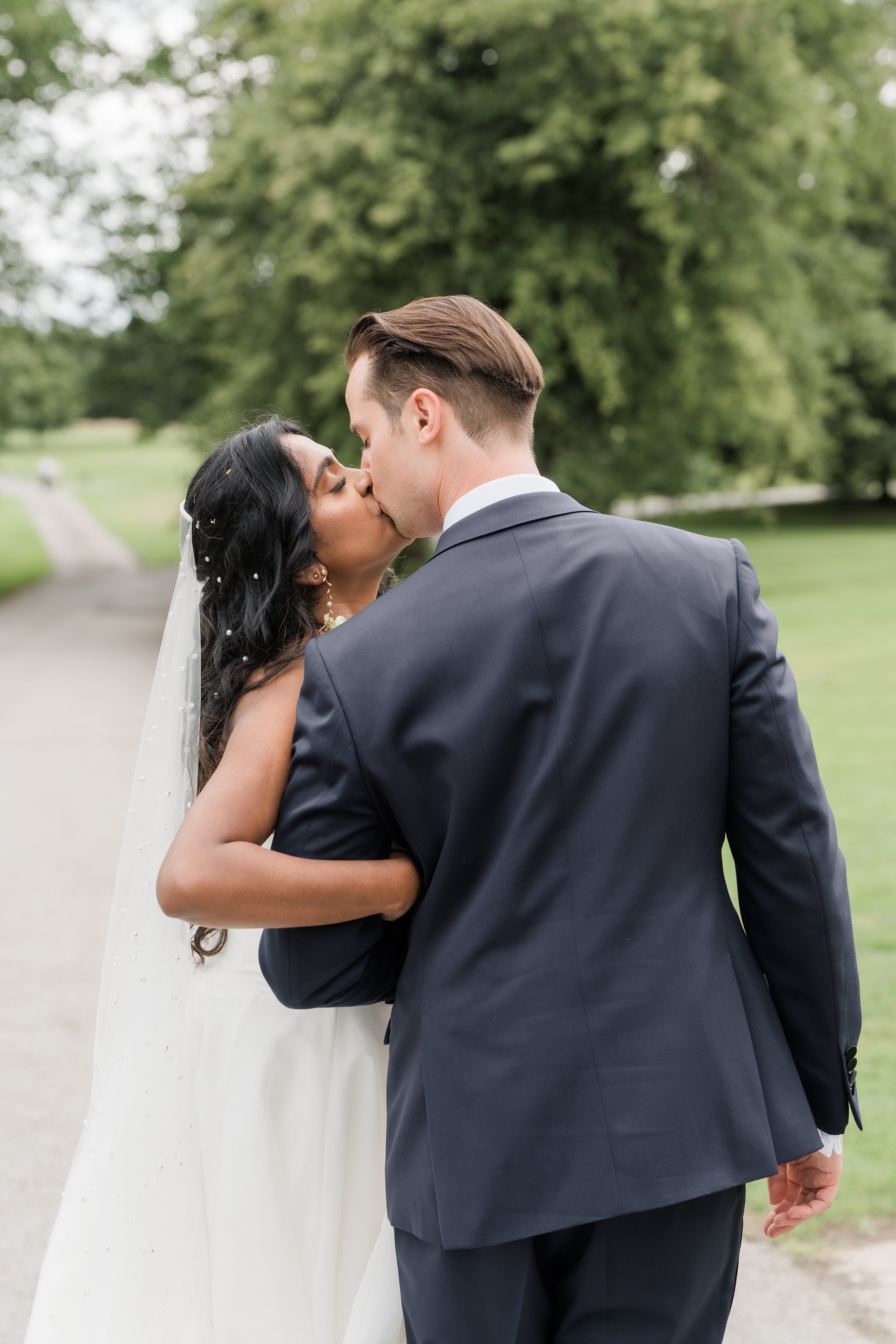 A bride and groom kiss outdoors on a wedding day, with trees and a grassy field in the background.