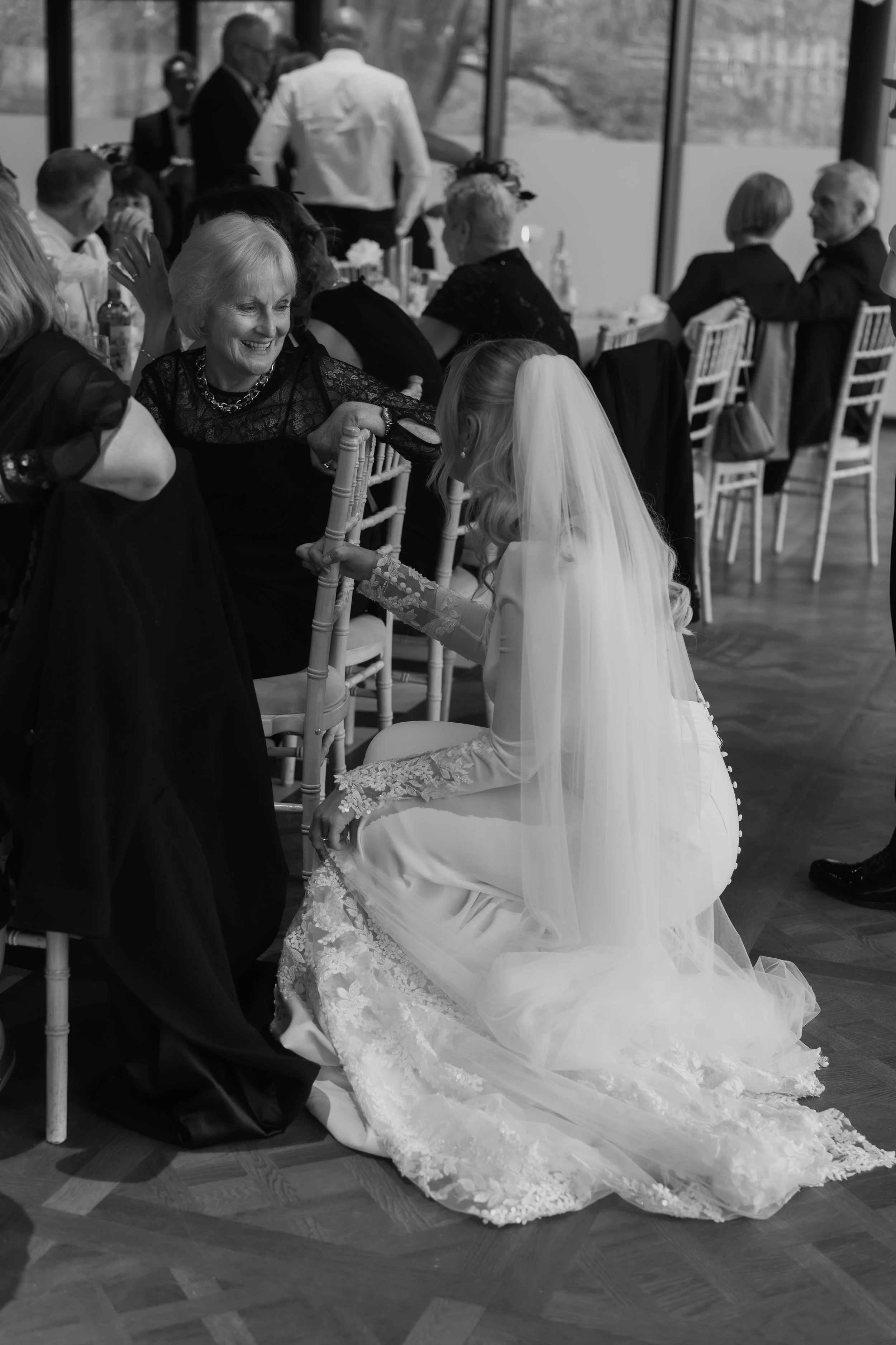 A bride in a wedding dress and veil kneels and holds hands with an elder woman, likely her grandmother, at a wedding reception. Other guests are seated at tables in the background.