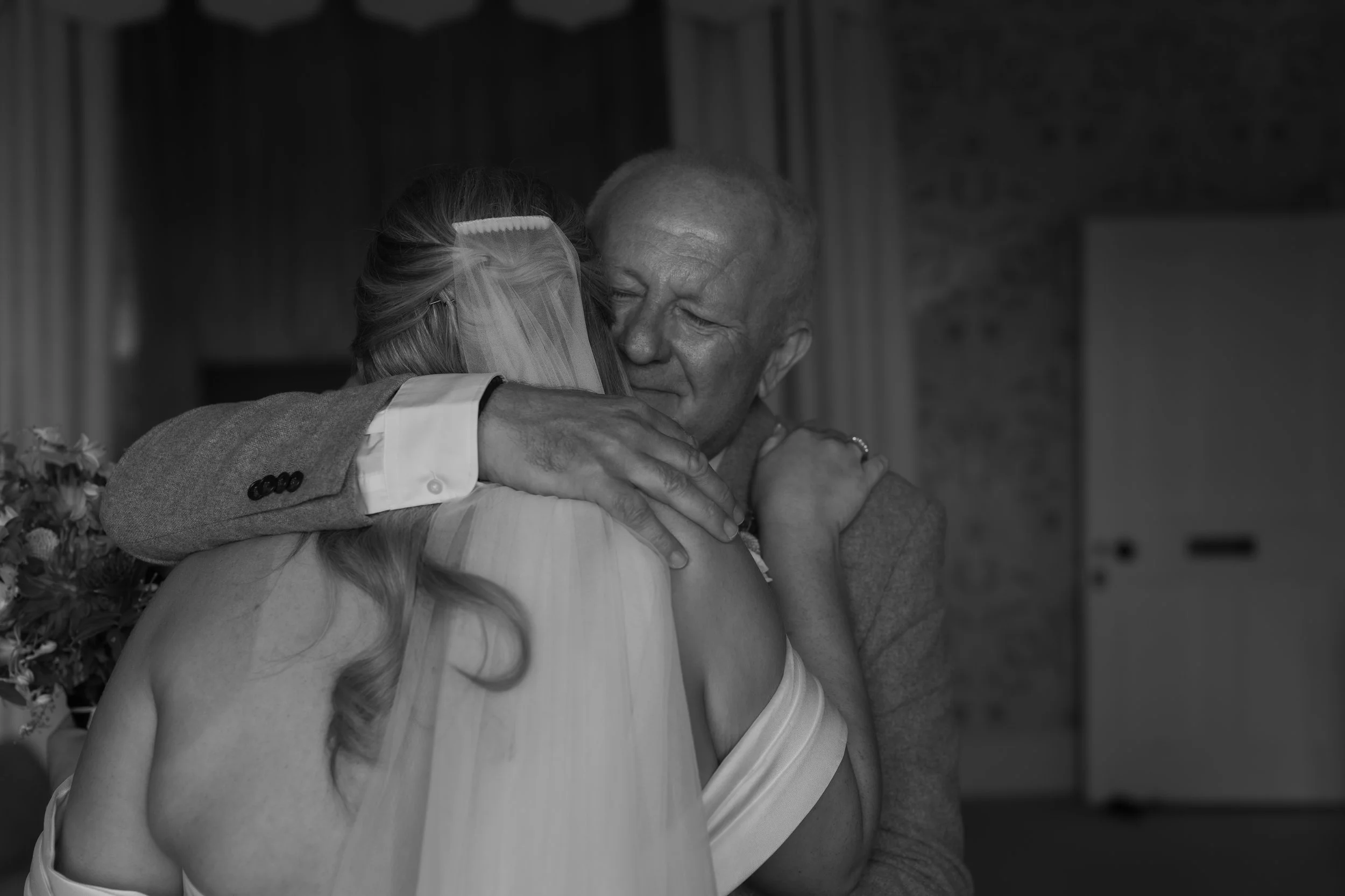 A heartfelt embrace between an elderly man and a woman in a wedding dress, sharing an emotional moment indoors.