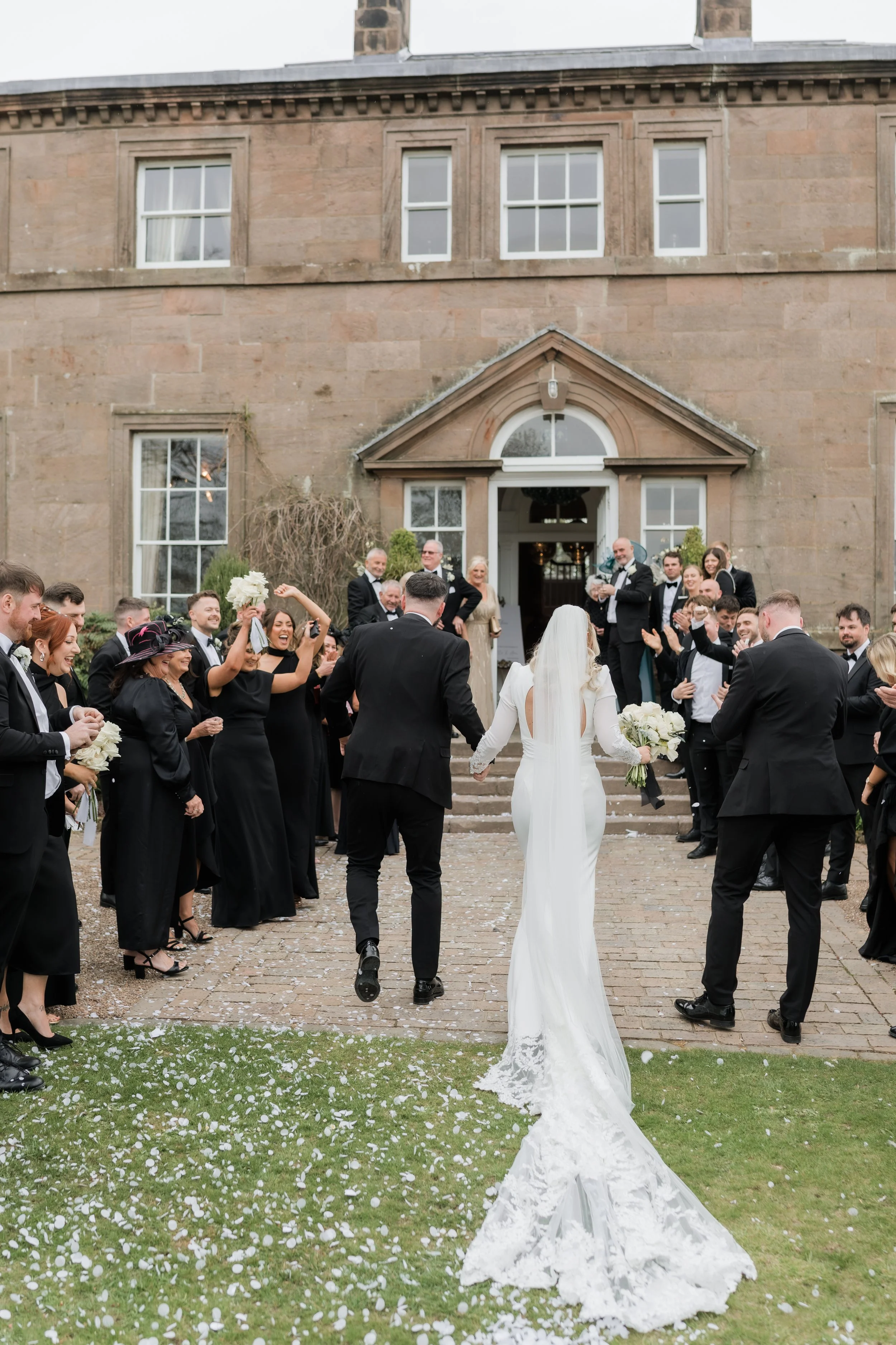 A bride and groom leave a wedding venue, walking hand-in-hand, followed by celebrating guests, outside a large stone building.