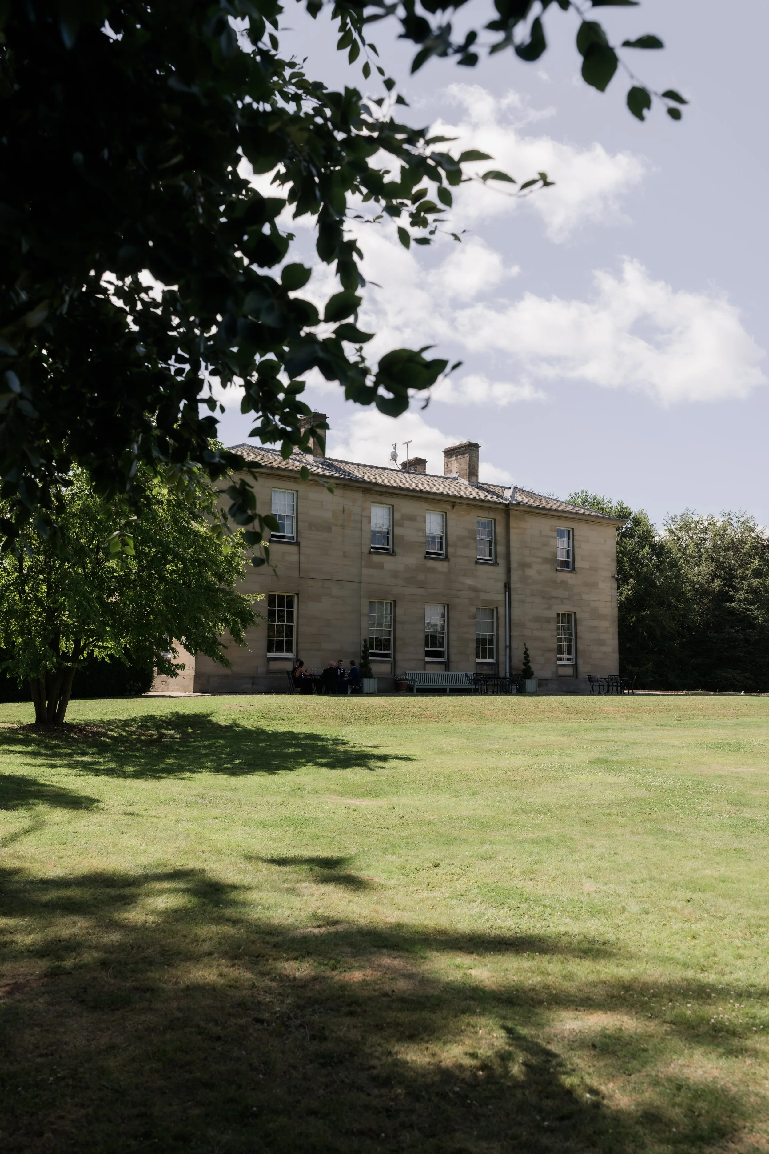 A park with well-maintained grass, large trees, and a building with multiple windows in the background. People are sitting on benches and chairs near the building, enjoying the outdoors. The sky is partly cloudy.
