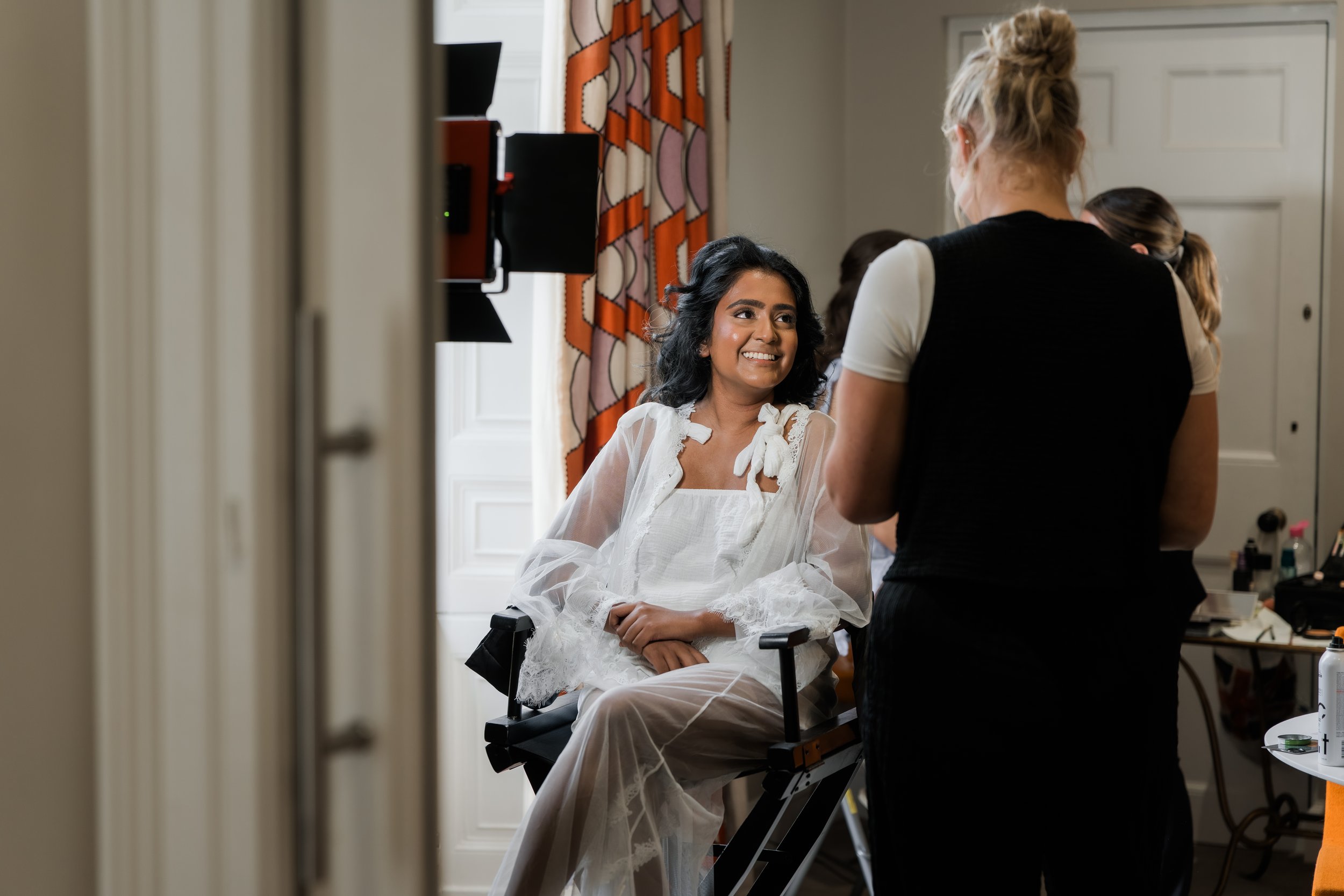 A woman sitting on a chair, getting her makeup done, smiling and looking at the makeup artist standing in front of her in a room with patterned curtains and a door.