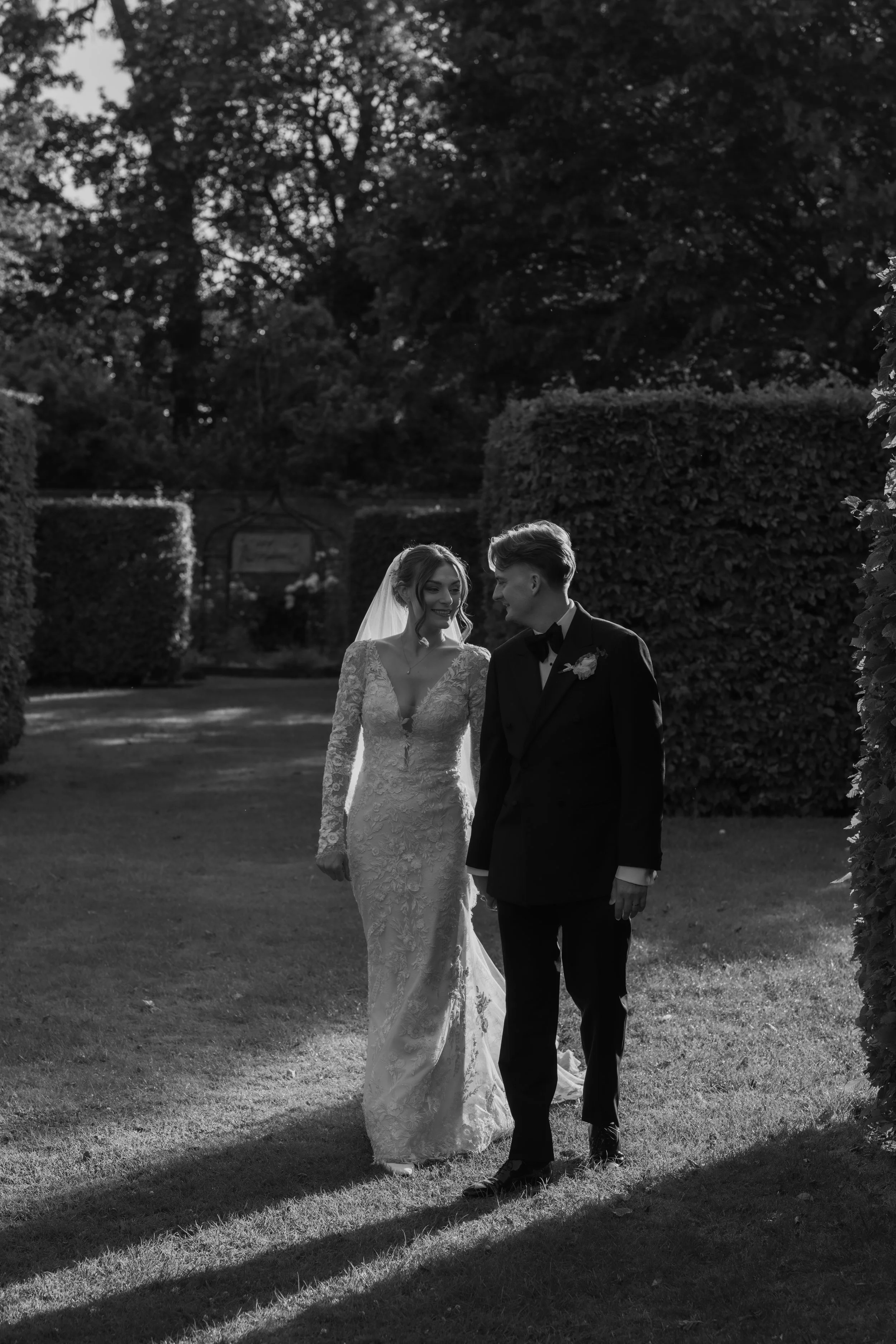 Black and white photograph of a bride and groom walking together outdoors on grass, surrounded by tall trees and hedges, with bright sunlight casting shadows.