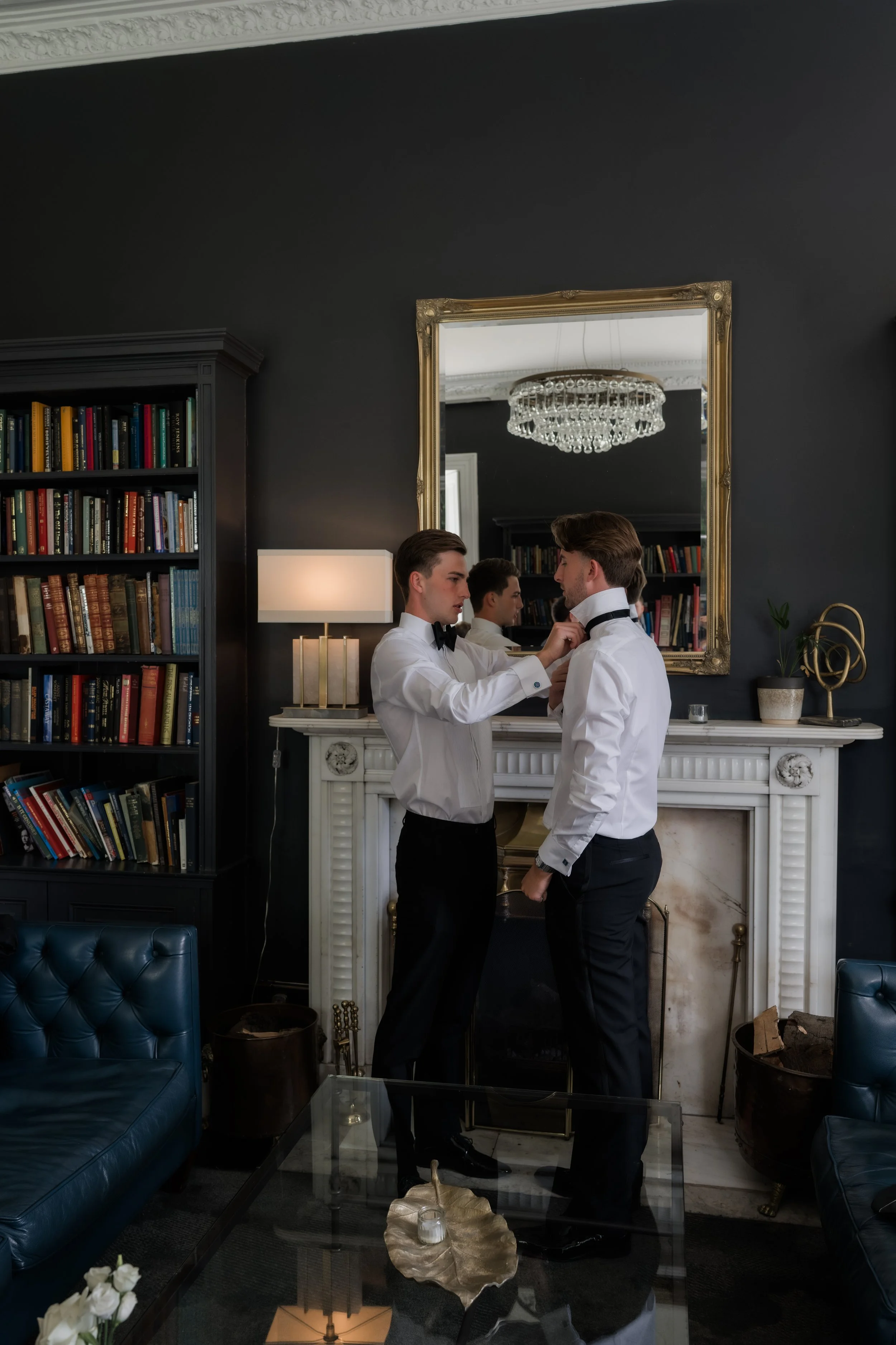 Two men dressed in formal attire standing close together in front of a large mirror in a room with dark walls, a white fireplace, bookshelf, and chandelier above.