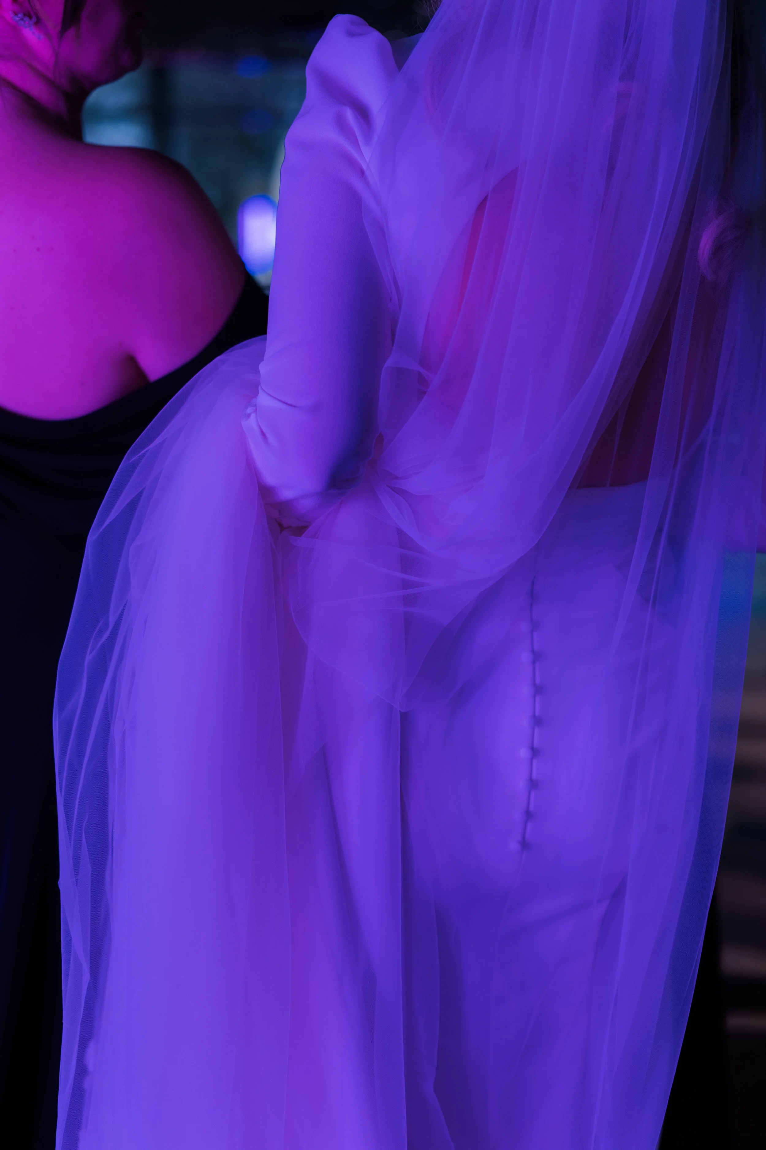 Close-up of two women in a dimly lit space wearing elegant outfits with blue and purple lighting. One woman is in a black off-the-shoulder dress, while the other wears a flowing, sheer, purple gown with buttons along the back.