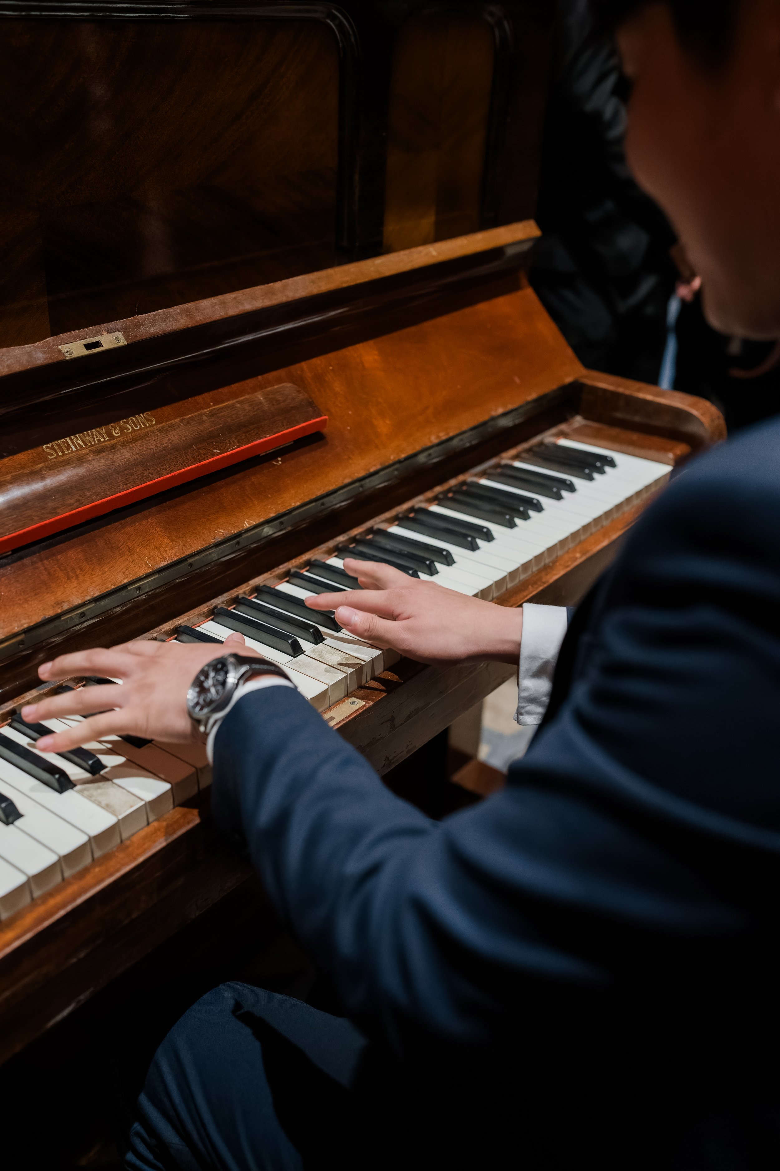 Person playing a vintage wooden upright piano, wearing a dark suit and a watch.
