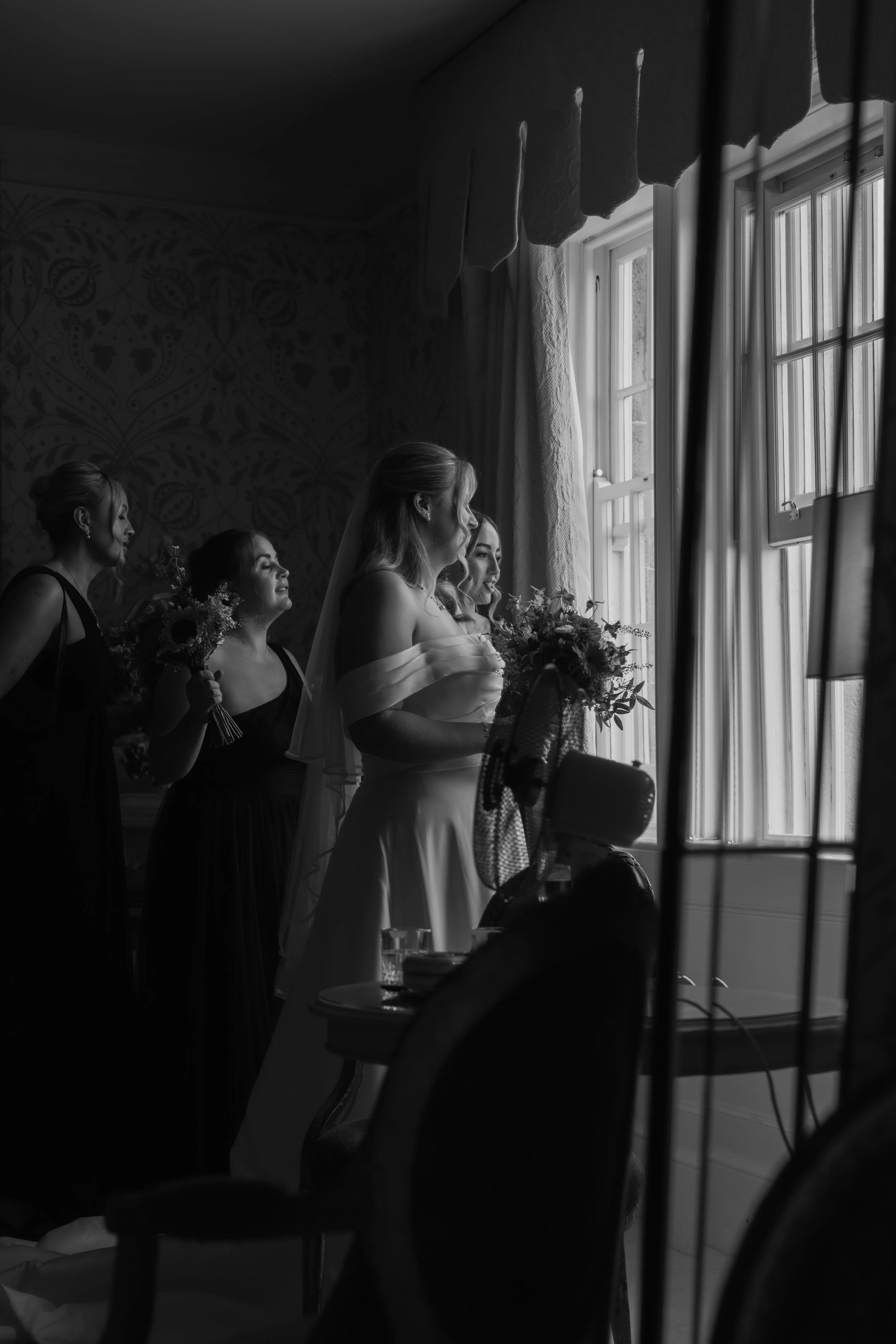 Black and white photo of a bride and her bridesmaids looking out a window before a wedding.