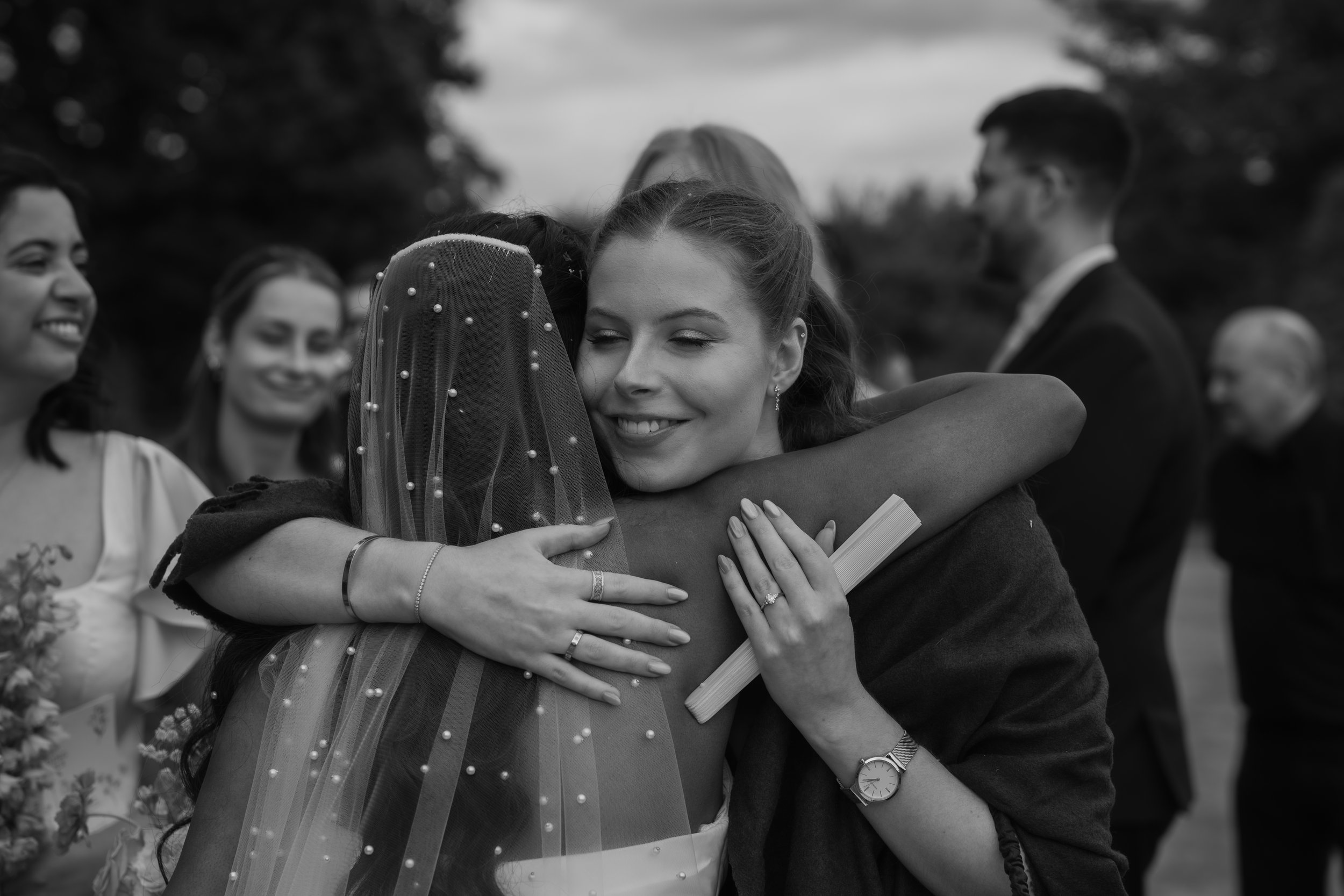 Two women hugging, one wearing a wedding veil, at a wedding celebration outdoors.