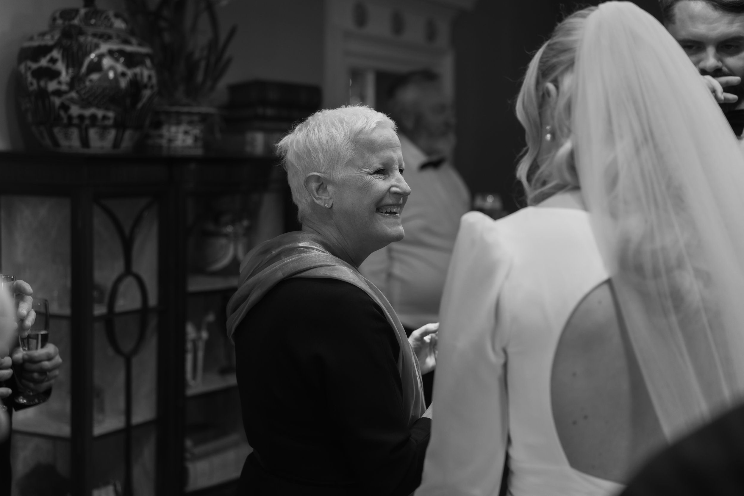 A woman with short white hair smiling and talking to a bride wearing a wedding dress and veil in an indoor setting.