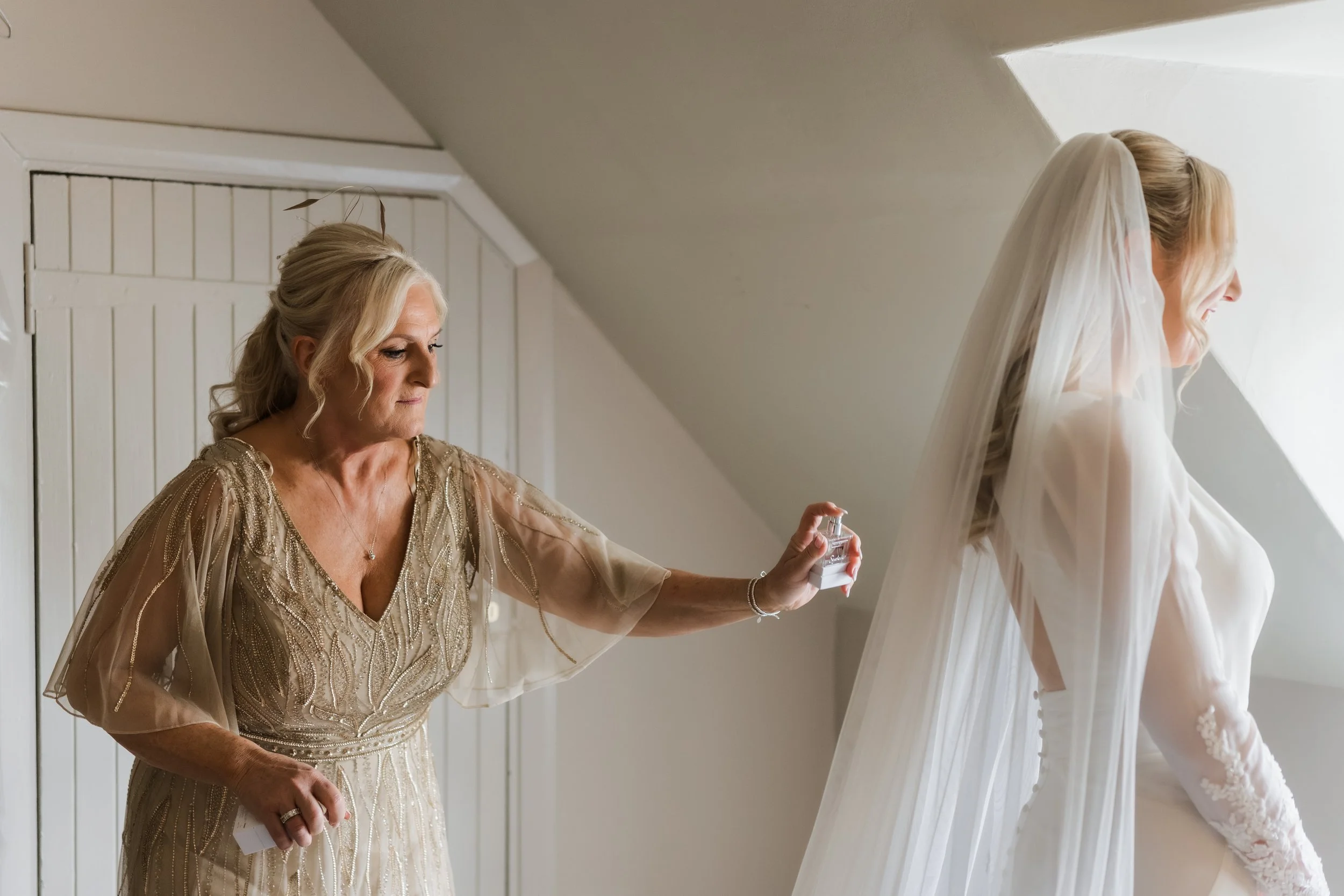 A bride smiling as her mother spritzes her with perfume in a room with sloped ceilings.
