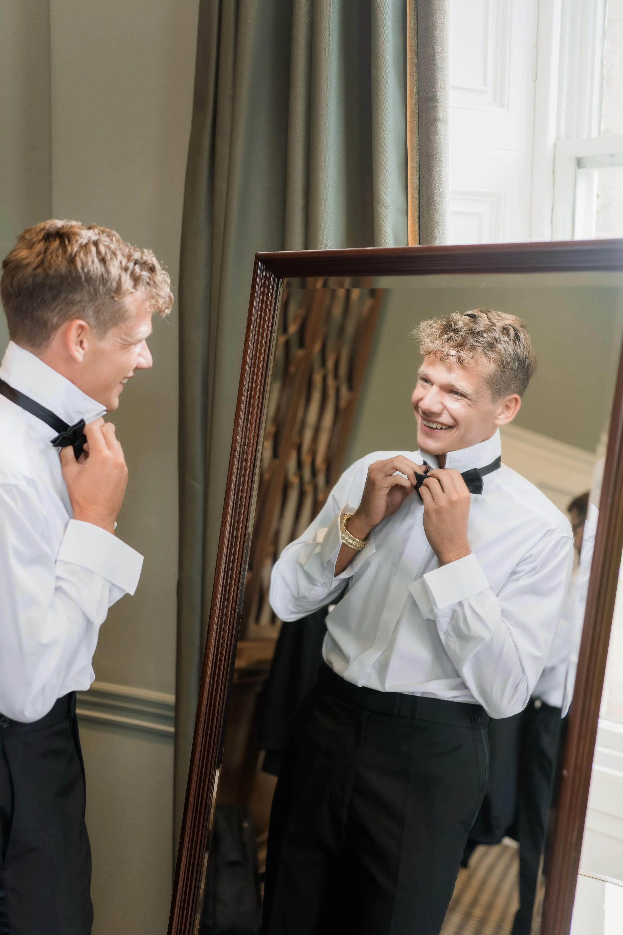 A man in formal attire adjusting his black bow tie while looking at himself in a large mirror. He is smiling and appears happy, with a green wall and window with curtains in the background.