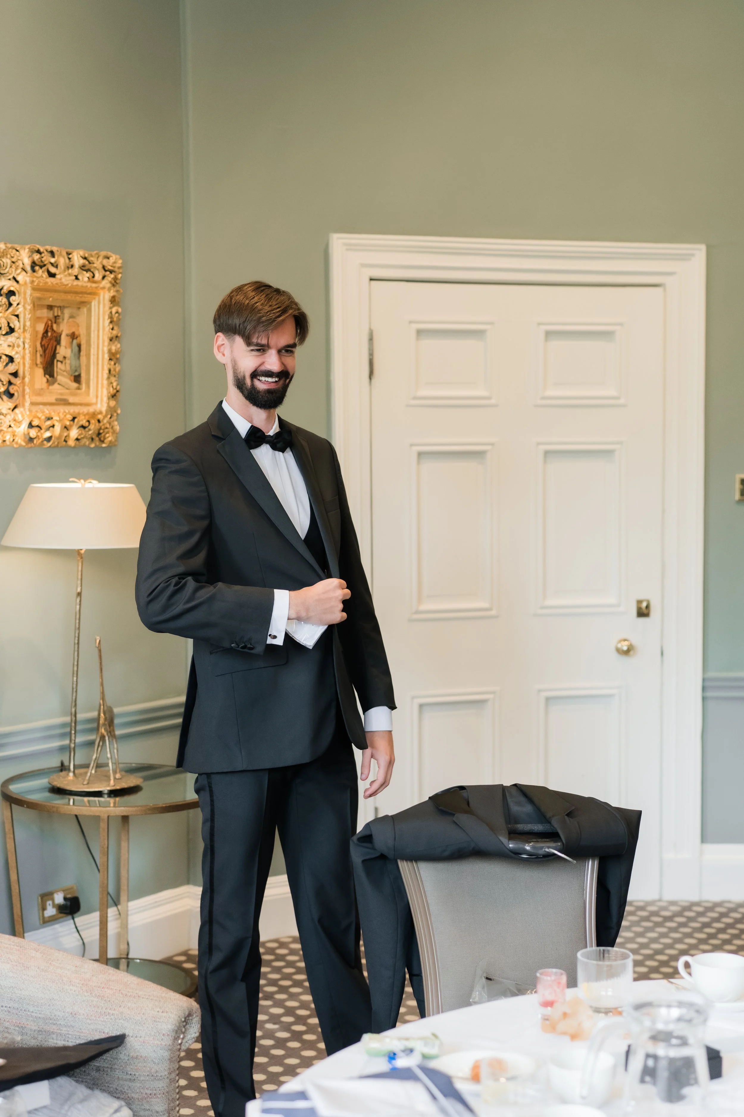 A man in a tuxedo with a bow tie smiling and adjusting his jacket in a room with a white door and wall art.
