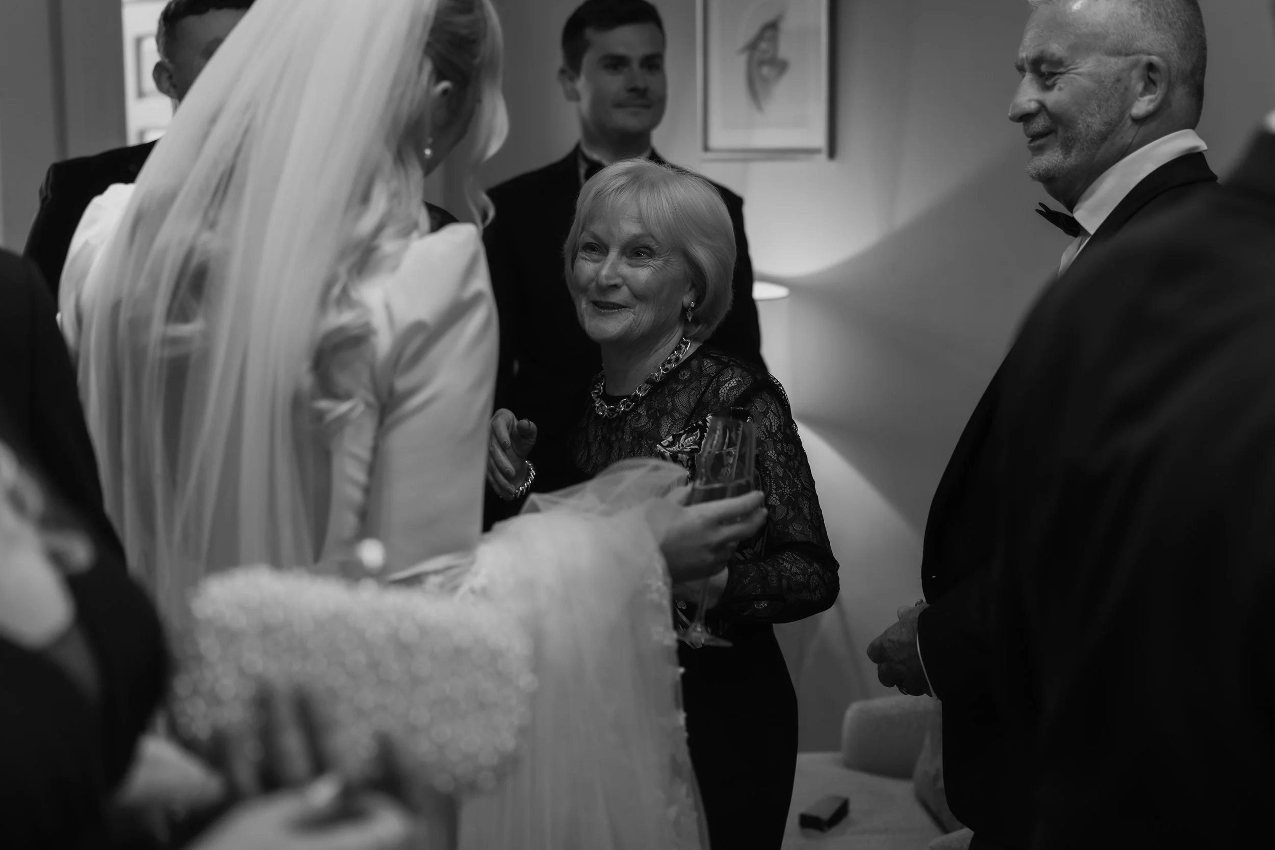 Black and white photo of a wedding reception with guests talking, some holding drinks, in a cozy indoor setting.
