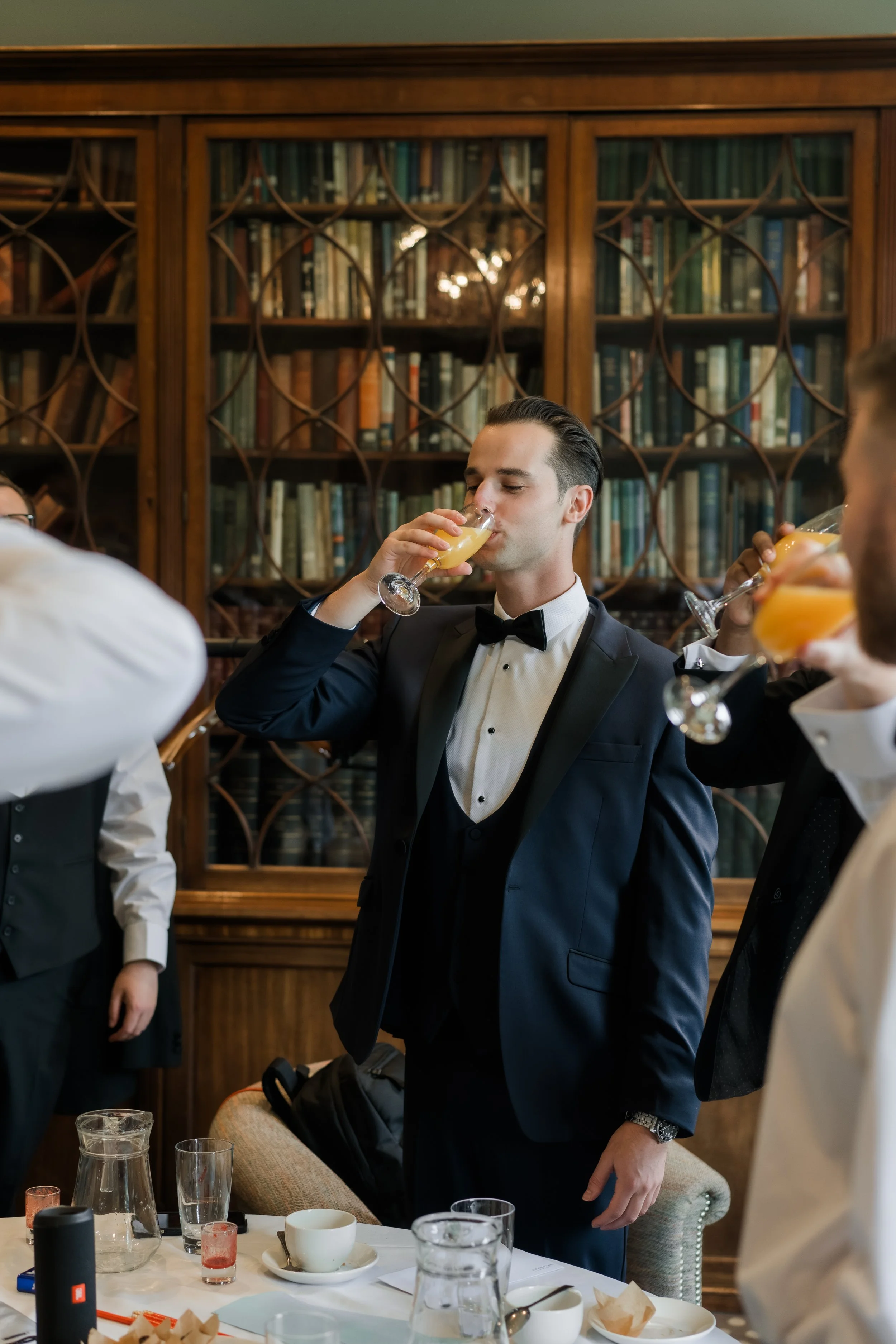 A man in a tuxedo and bow tie is drinking a mimosa at a formal gathering in a room with wooden bookshelves filled with books.