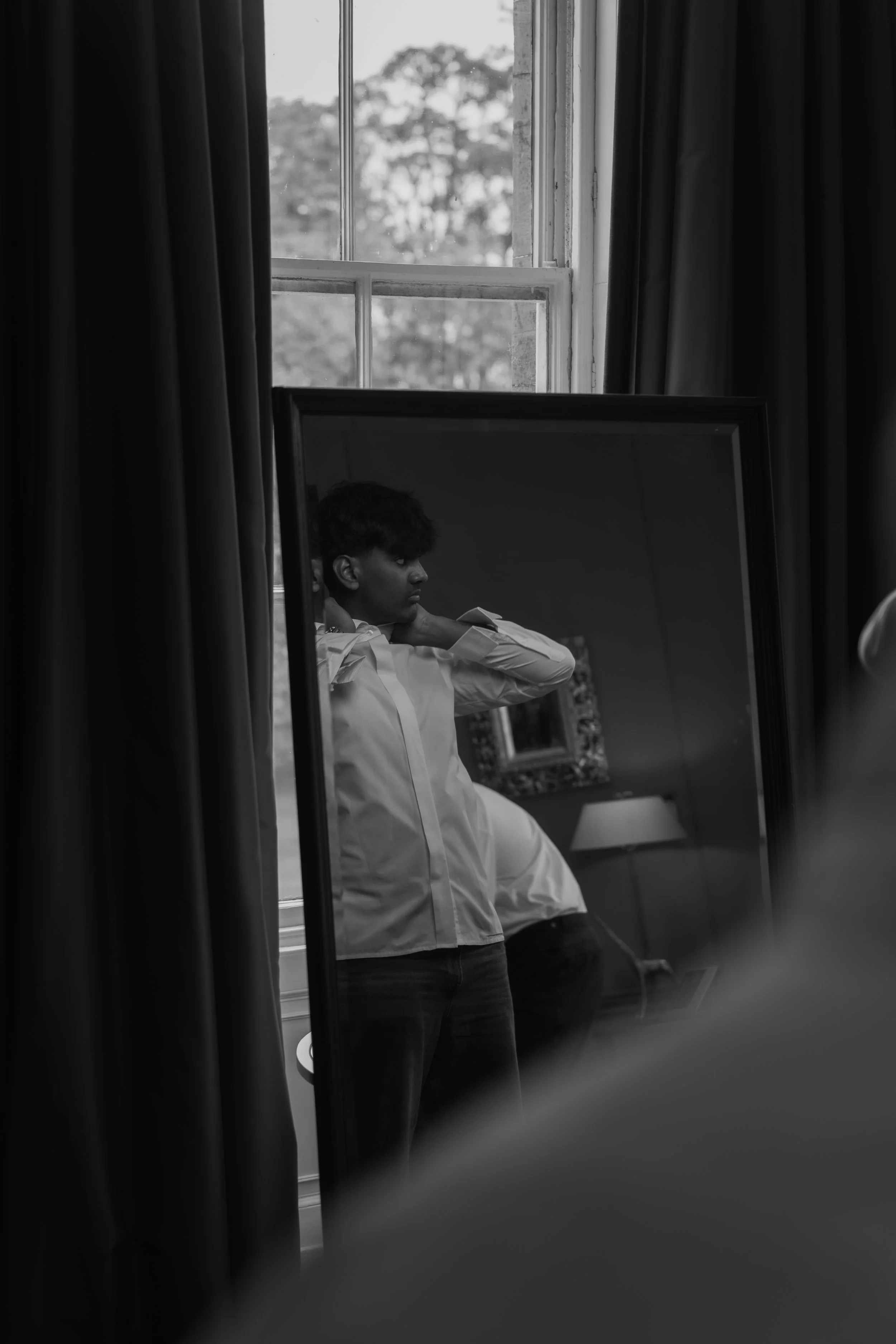 A young man stands in front of a mirror adjusting his shirt, reflected in a black-and-white photograph taken in a room with a window and curtains.