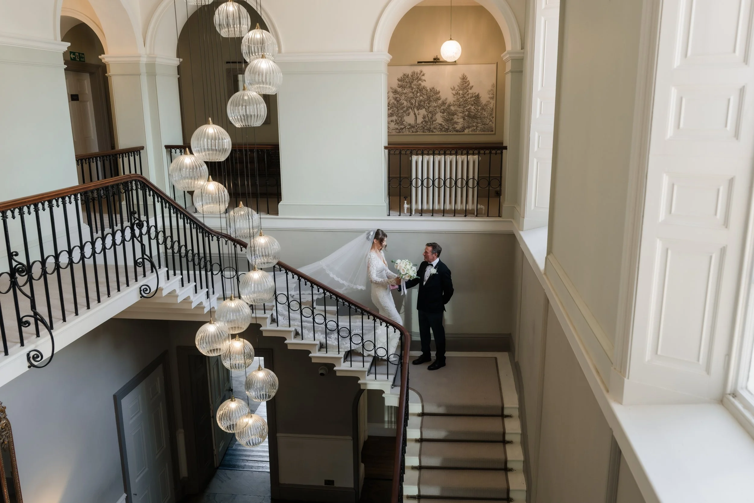 A bride and groom standing on staircase inside a building with high arch ceilings, chandelier, and wall art of trees.