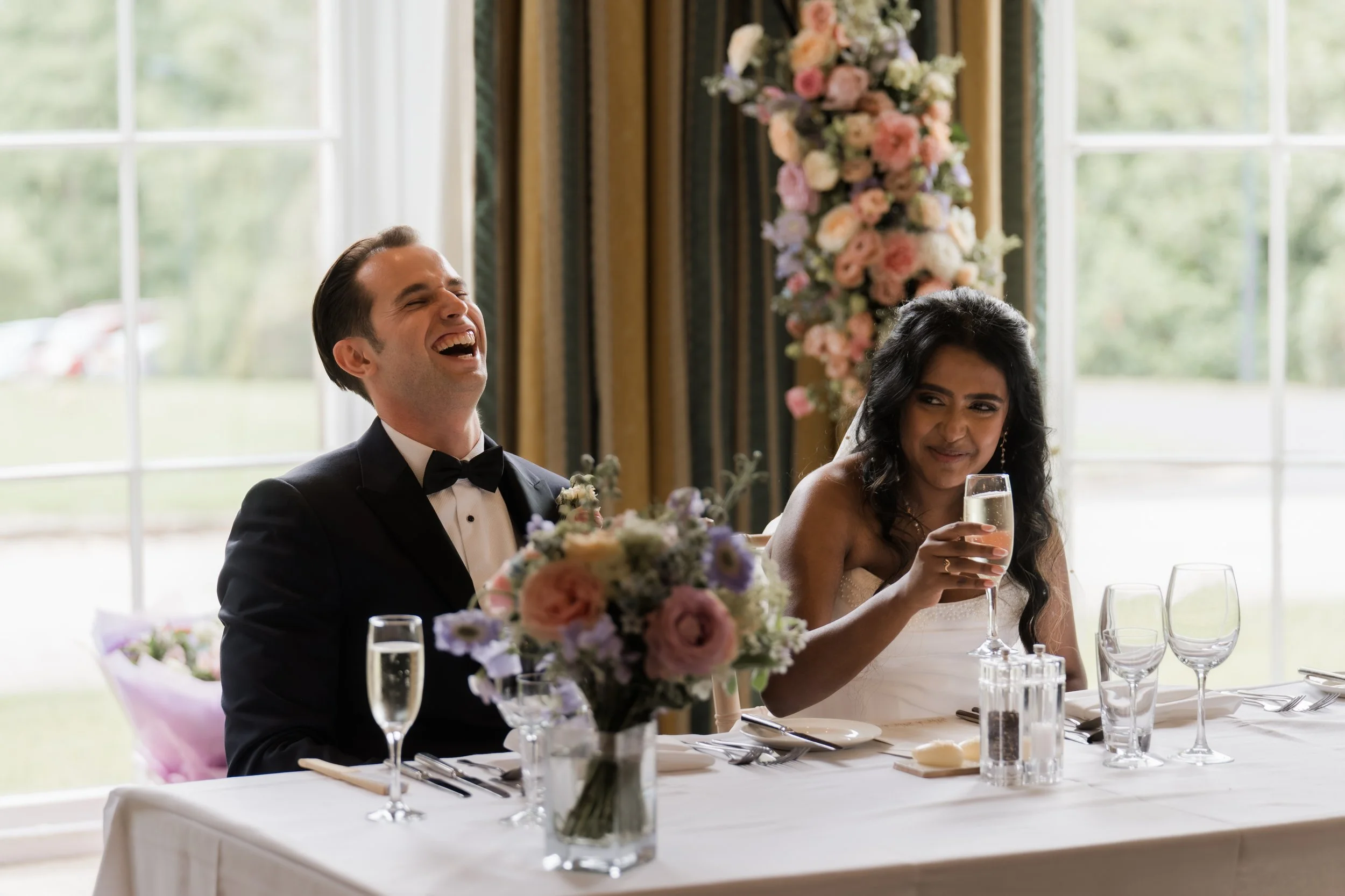 A bride and groom sitting at a wedding reception table, the groom laughing and the bride looking amused while holding a glass of champagne, with floral decorations and large windows in the background.