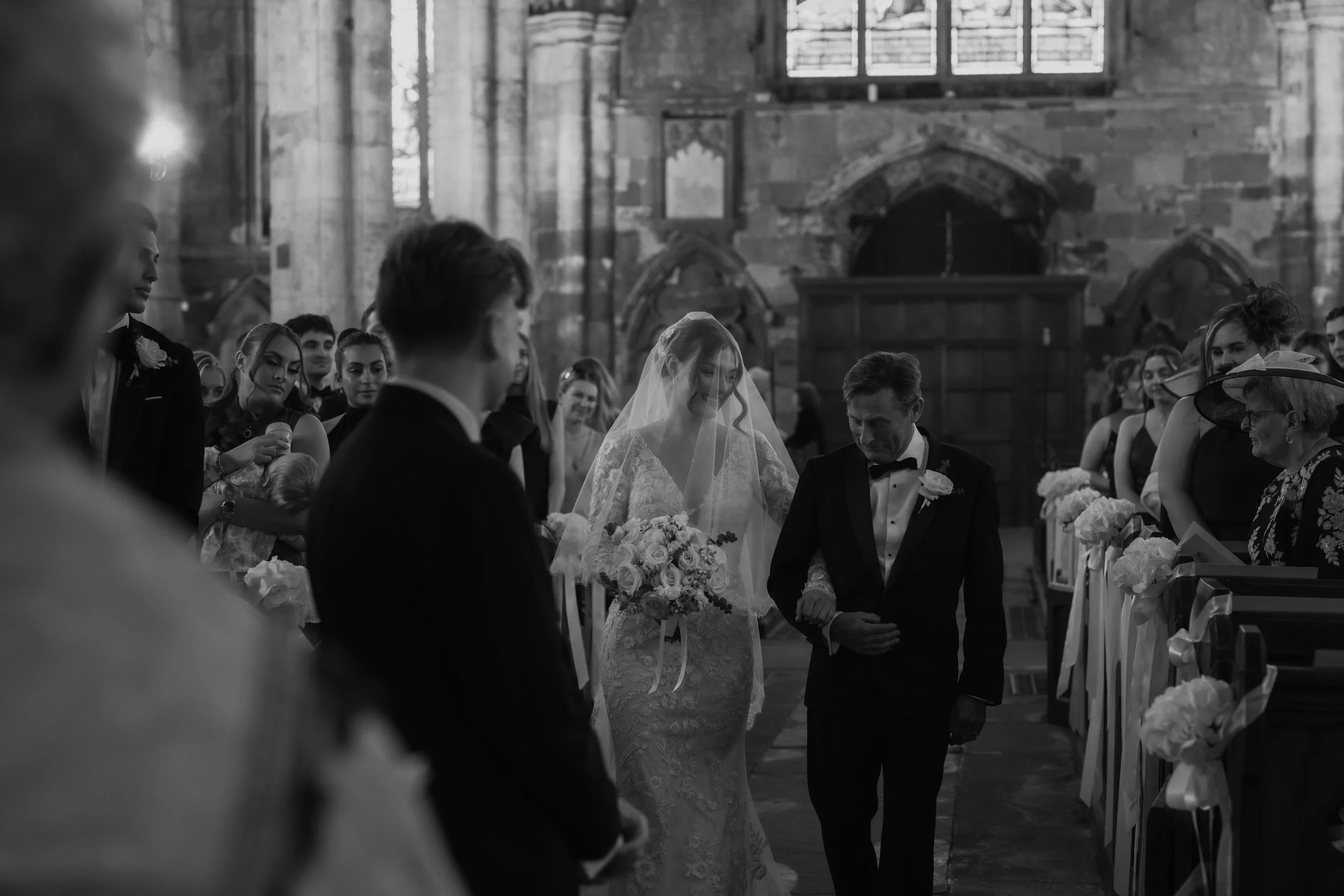 Black-and-white photo of a wedding ceremony inside a church, with the bride and groom holding hands and surrounded by guests, some wearing formal attire and hats.