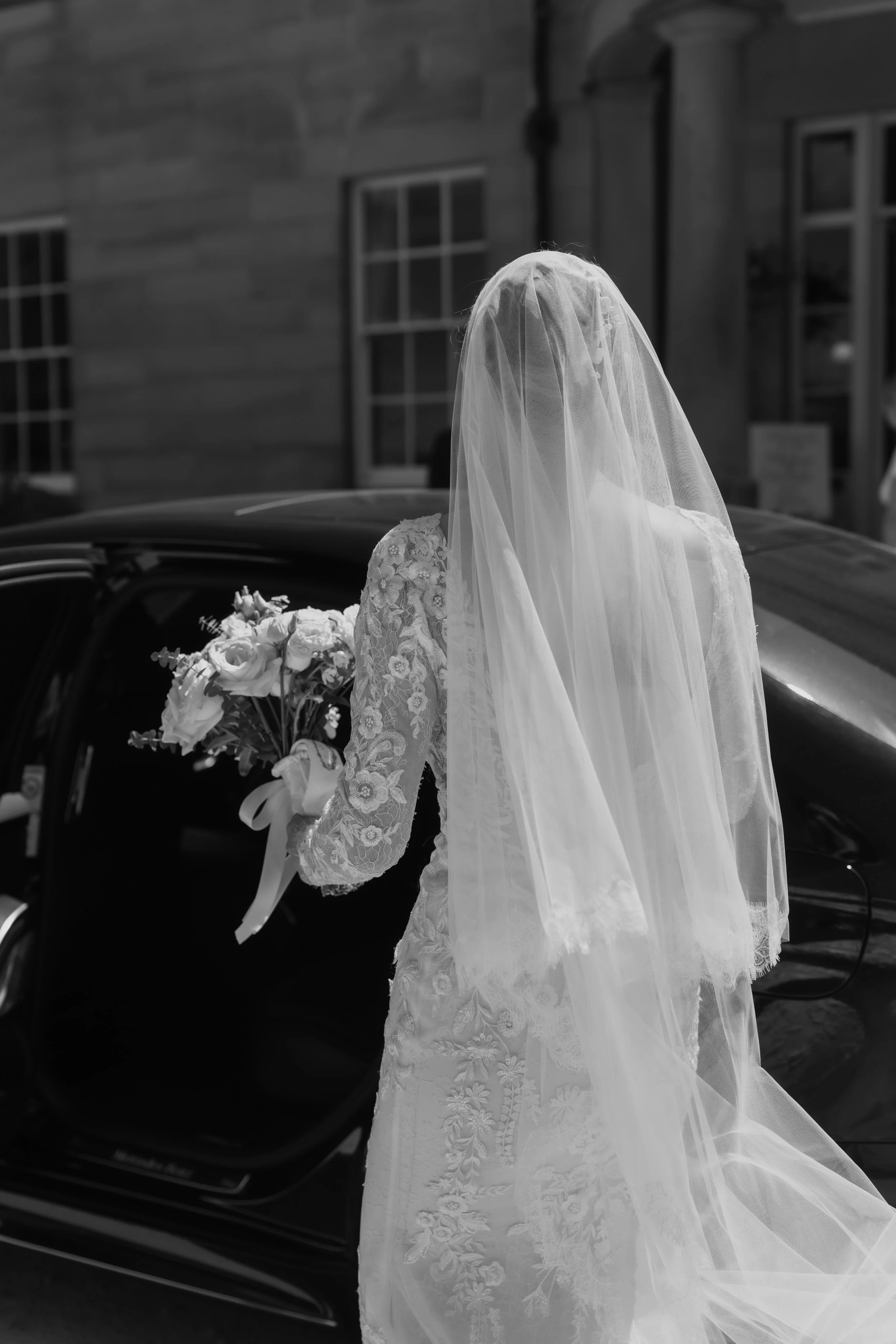 A bride in a lace wedding dress with long sleeves and floral embroidery, wearing a veil, holding a bouquet of white flowers, standing outdoors near a car and a house in the background.