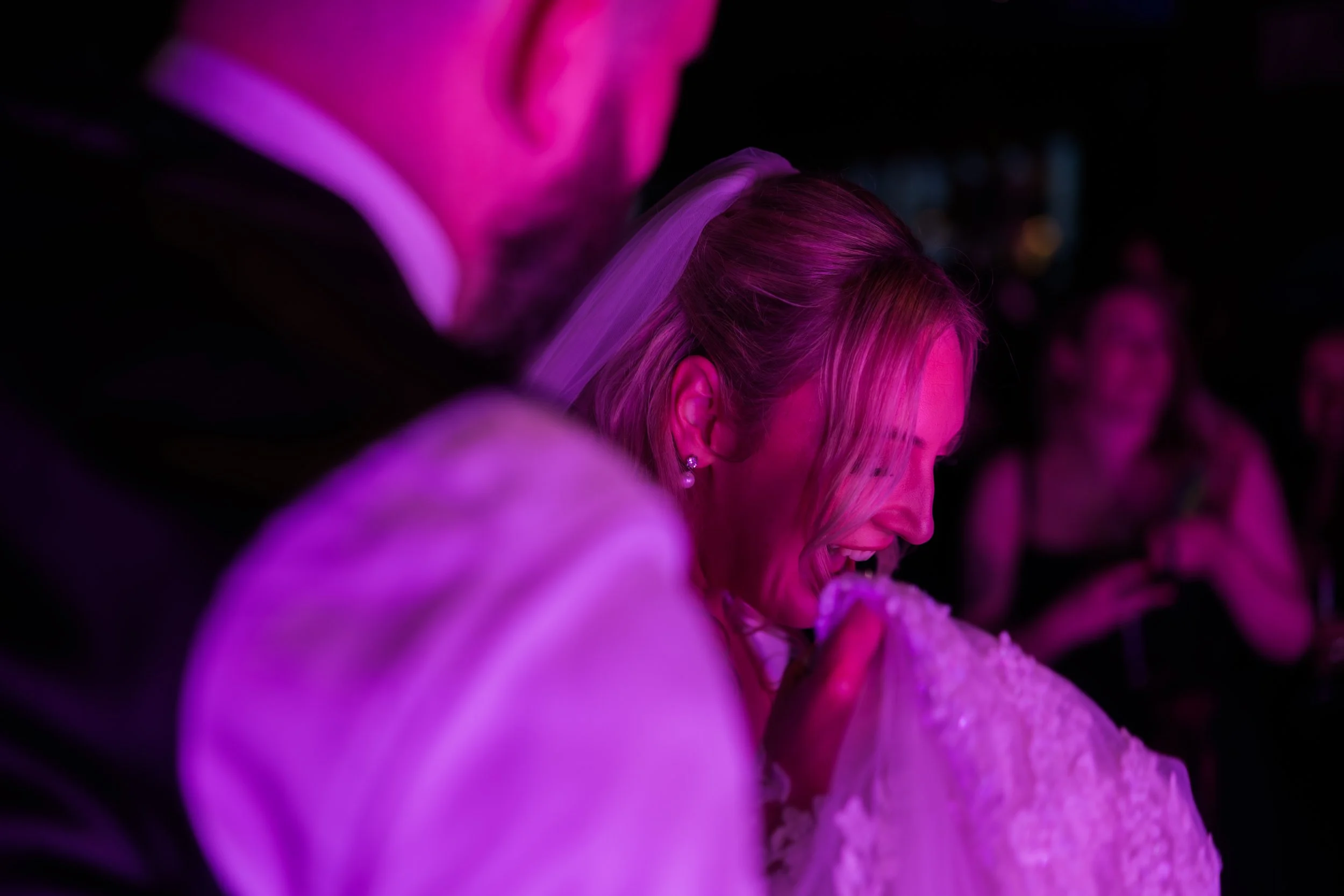 A woman with blonde hair and pearl earrings appears to be biting or eating a piece of cake at a celebration, with people in the background under pink lighting.