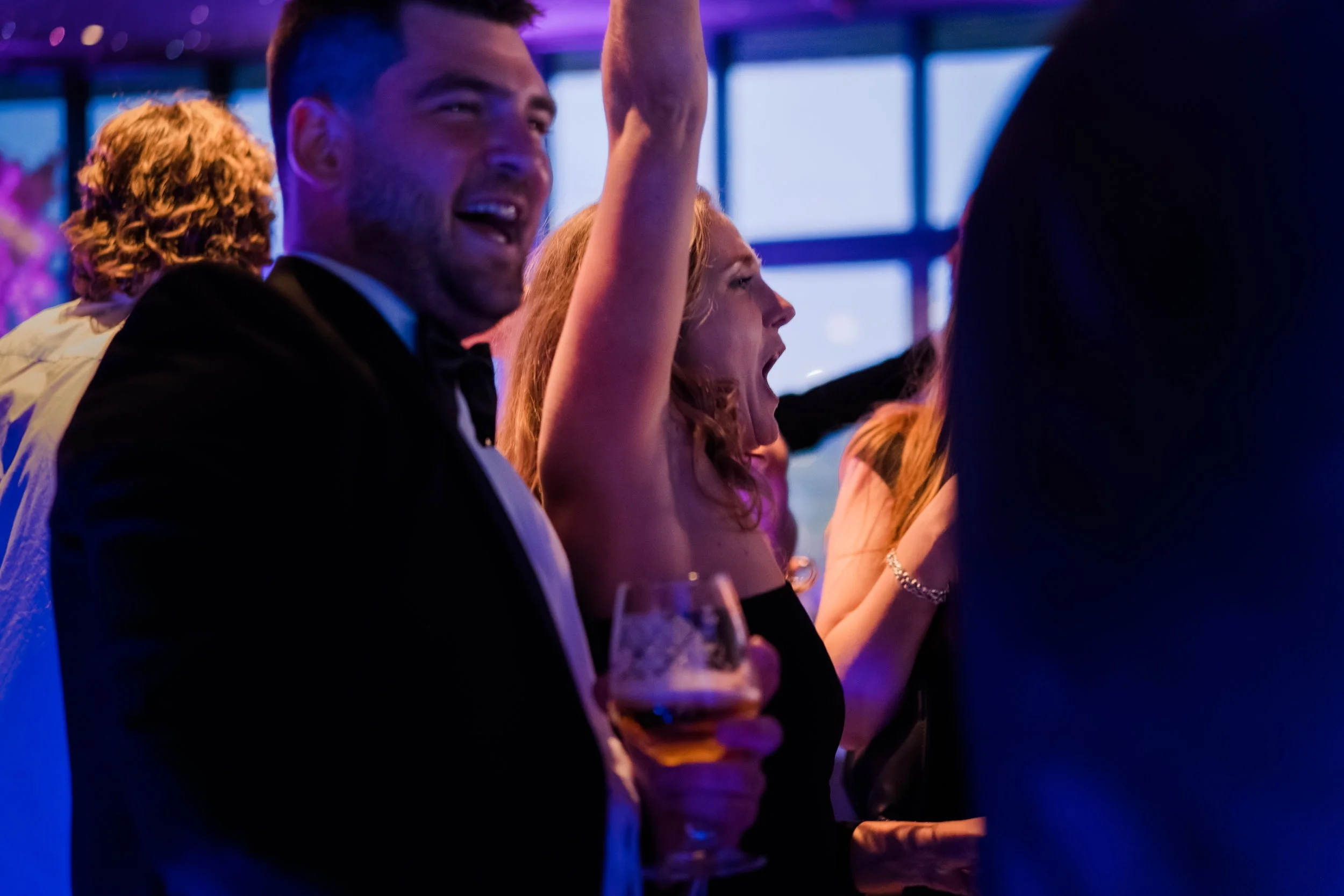 A group of people celebrating at a party or event, some with drinks in hand, in a lively setting with colorful lighting.