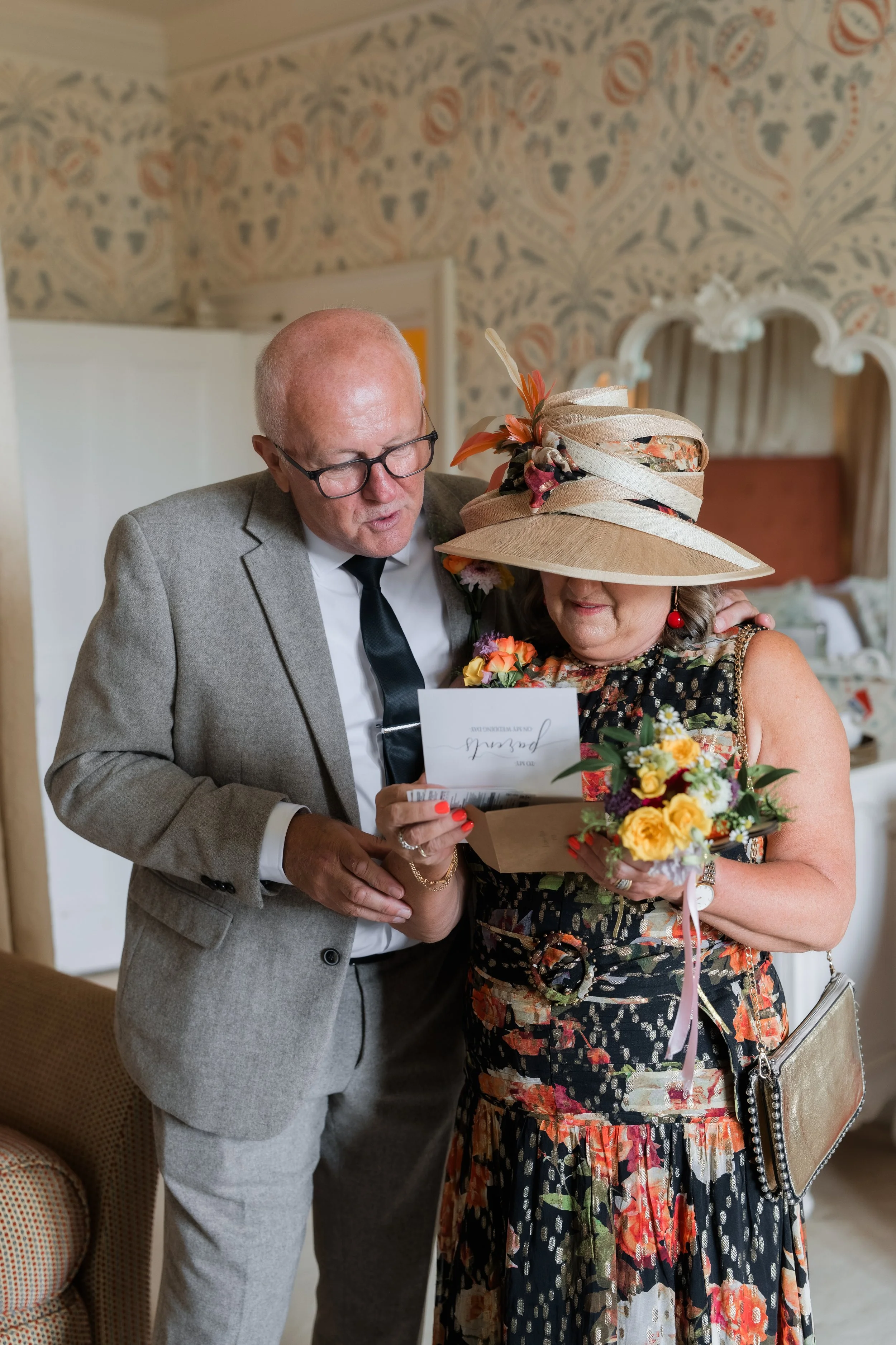 A man in a gray suit and a woman in a floral dress with a large hat, both looking at a card or note. The woman holds a bouquet of yellow and purple flowers.
