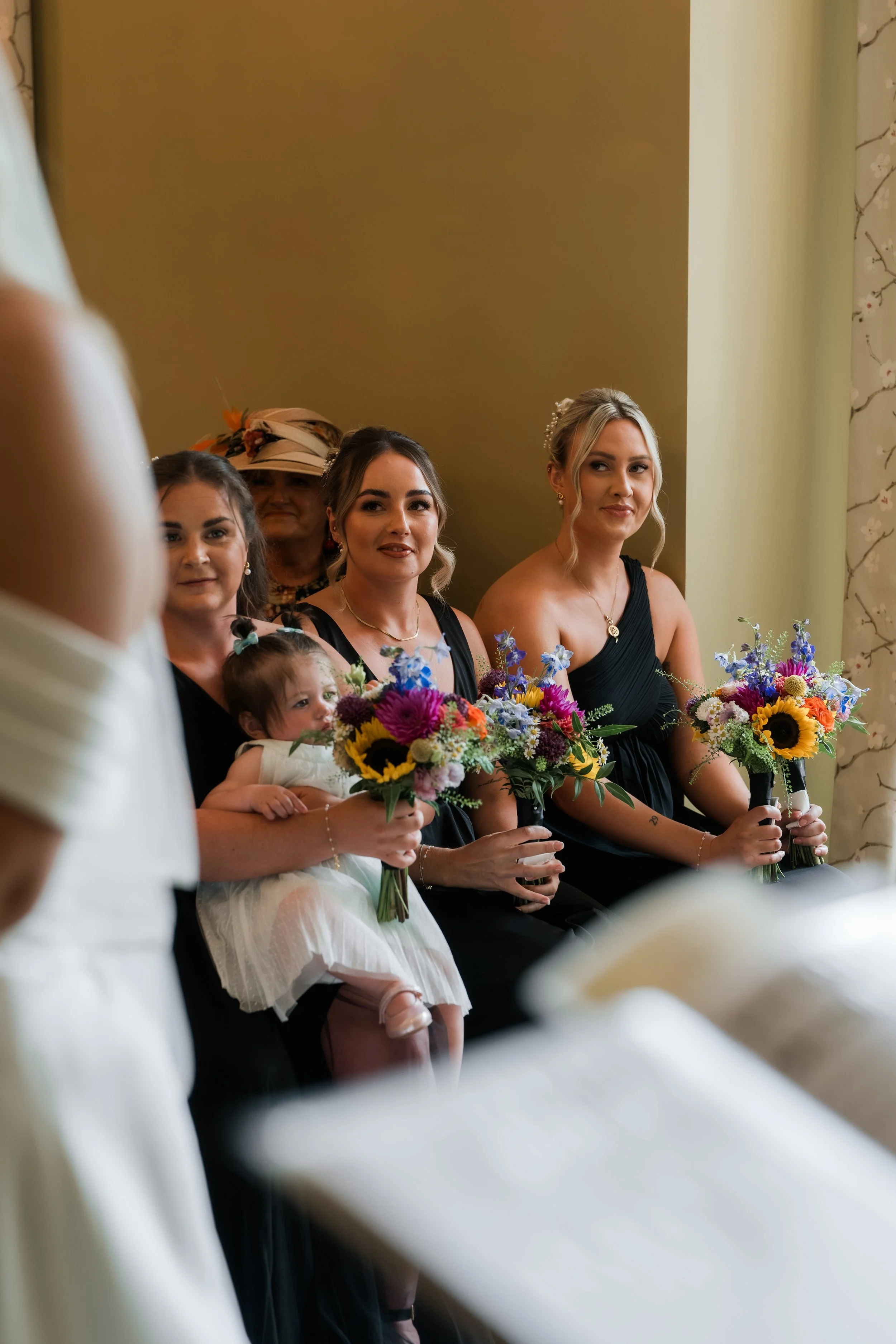 Bridesmaids sitting with bouquets of colorful flowers during a wedding ceremony.