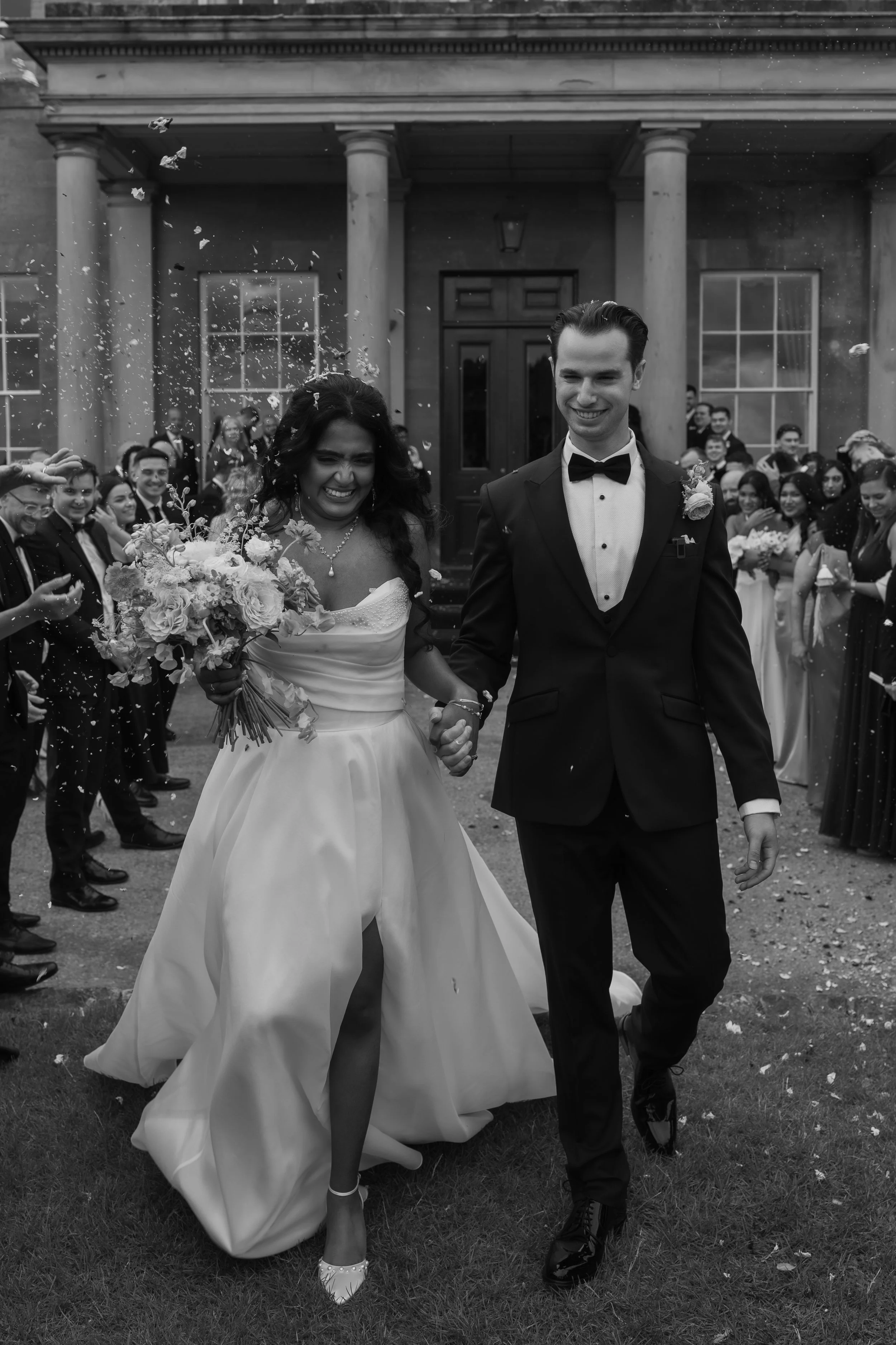A bride and groom walking hand-in-hand outside a building, smiling, surrounded by friends and family, with confetti falling around them.