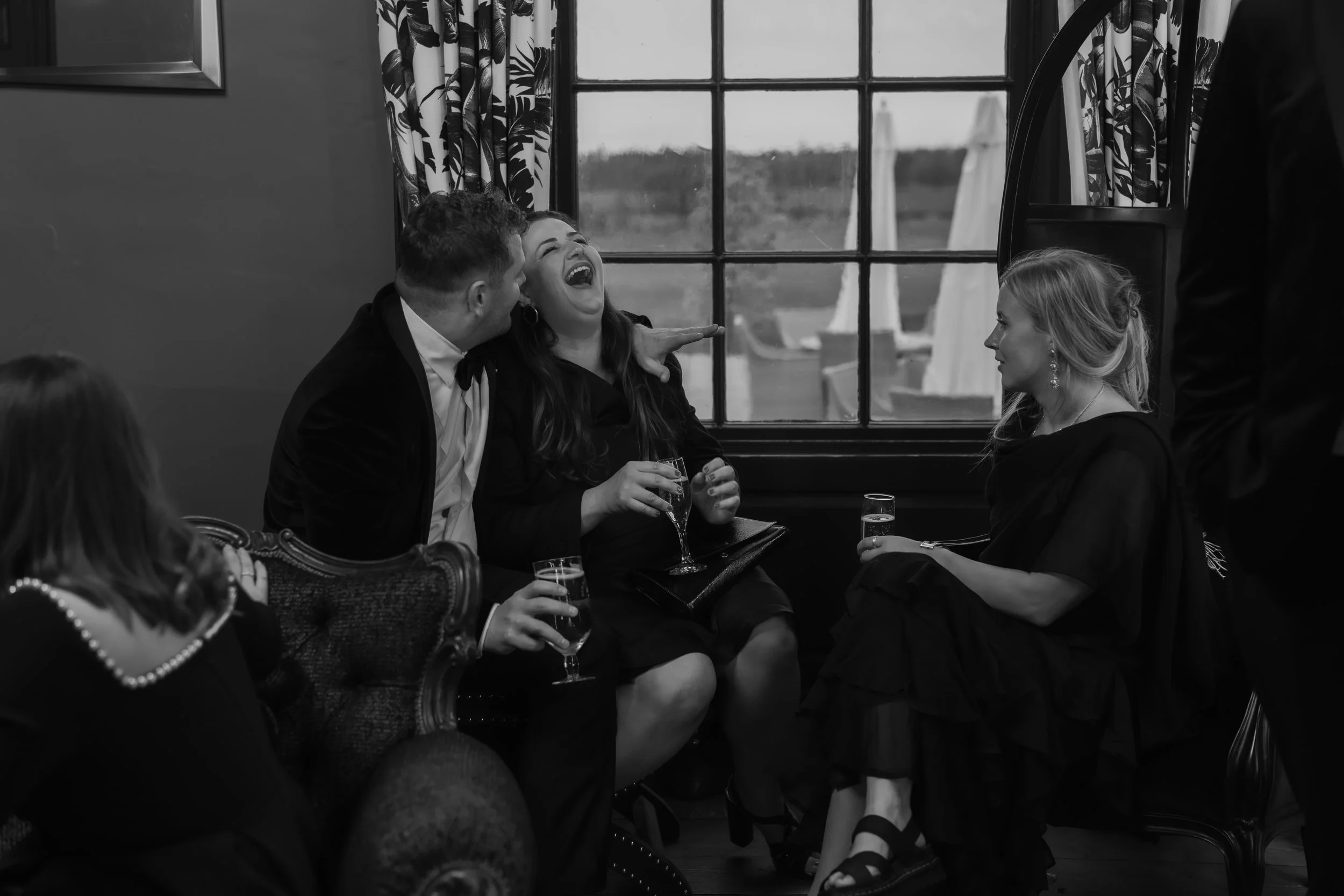 People gathered indoors, laughing and talking, with drinks in hand, in a cozy room with a large window and patterned curtains.