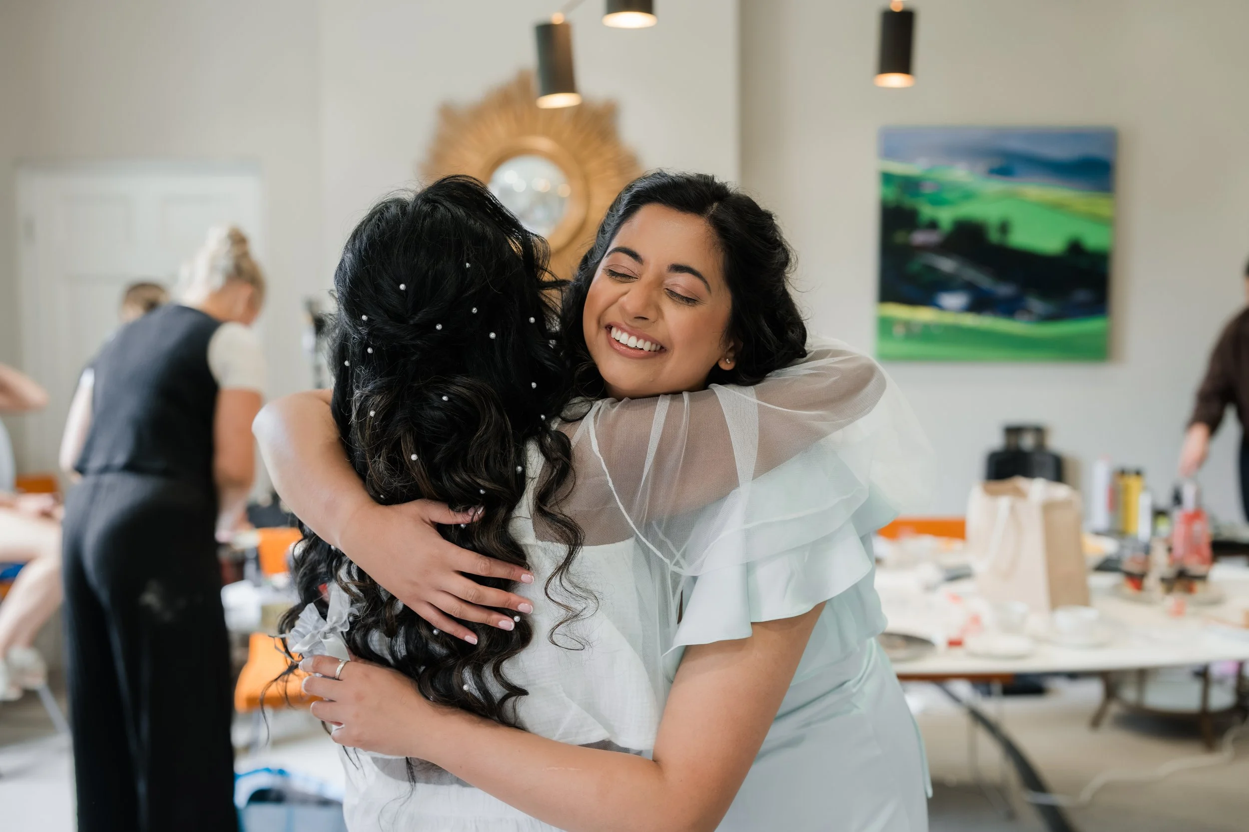 Two women hugging, one in a white dress, in a room with other people, makeup, and hair supplies.