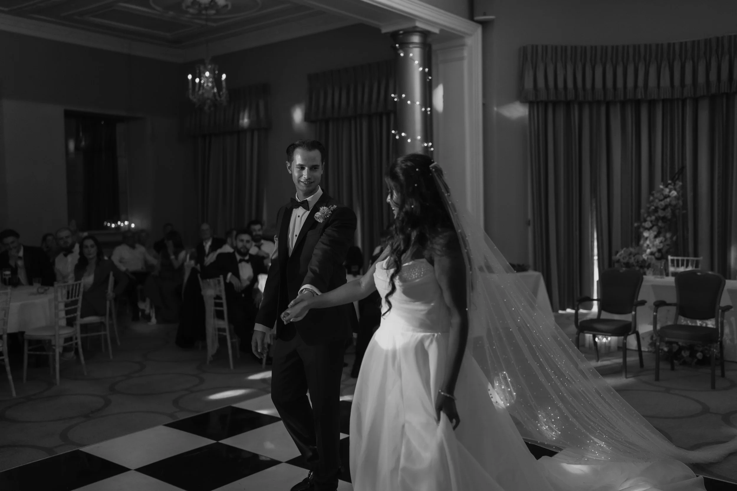 Black and white photo of a newlywed couple dancing at their wedding reception, with wedding guests seated at tables in the background.