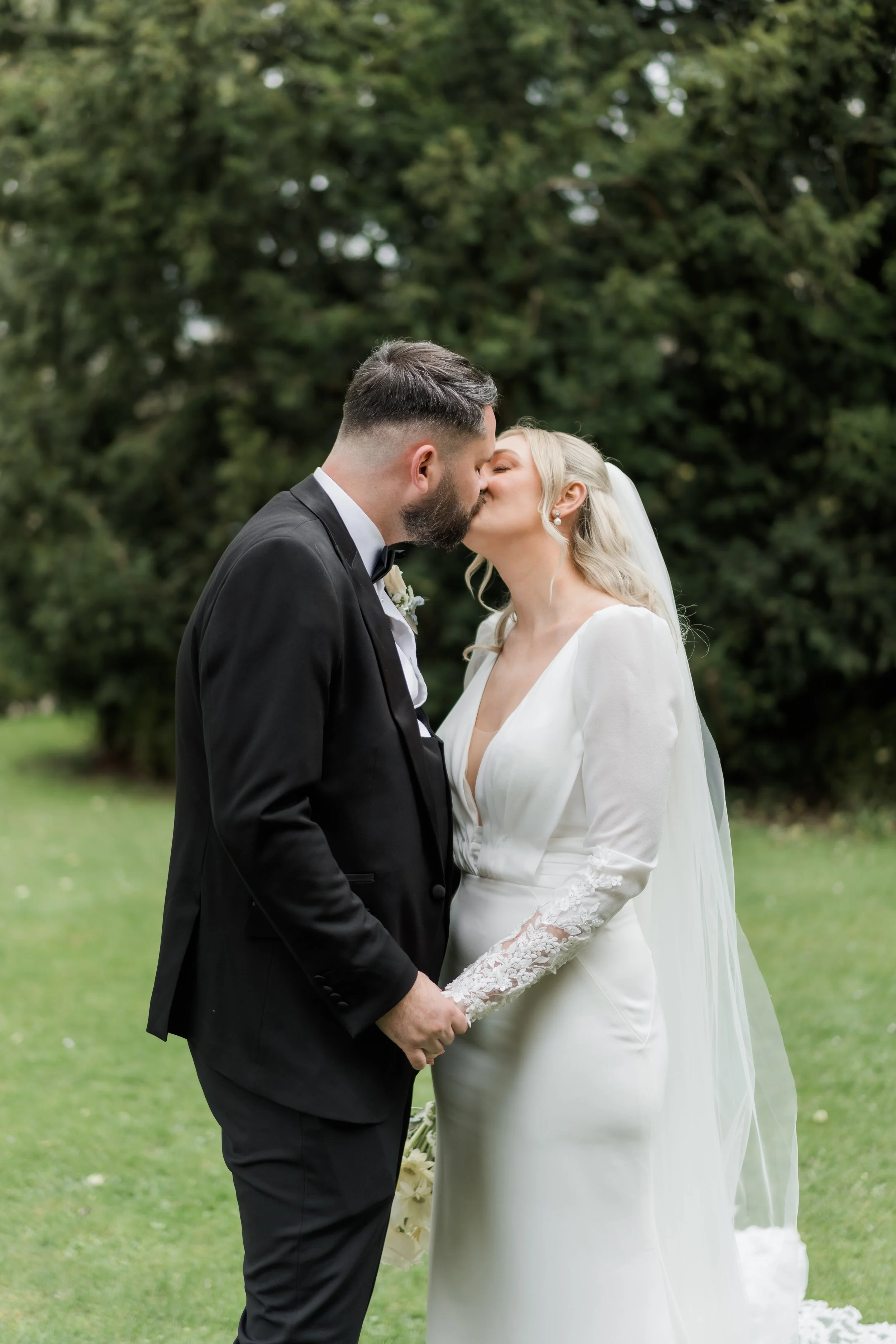 A bride and groom kissing outdoors on their wedding day, holding hands, with green trees in the background.