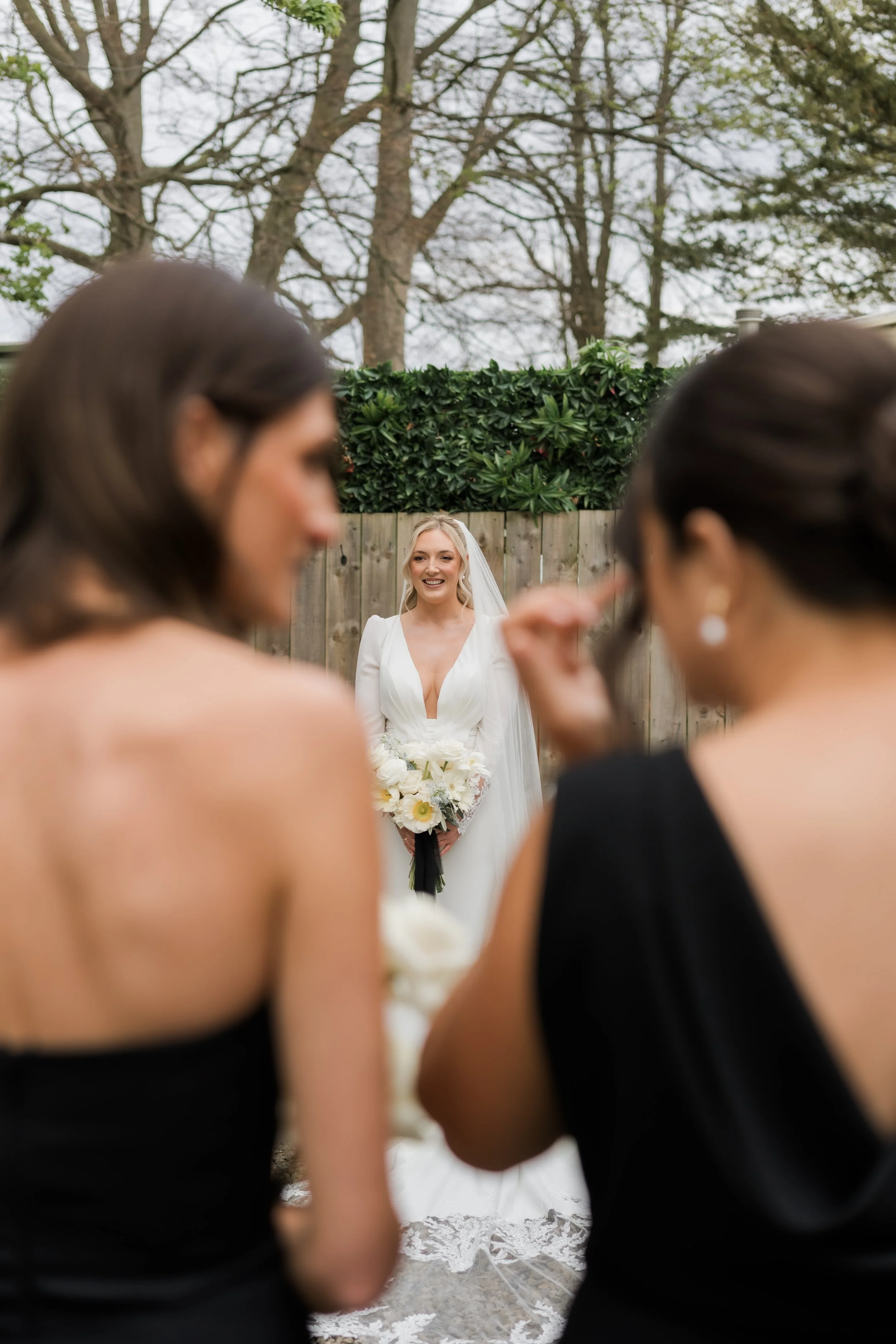 A bride with blonde hair in a white wedding gown holding a bouquet of white flowers, smiling as two women in black dresses prepare for her photo outside with a wooden fence and trees in the background.